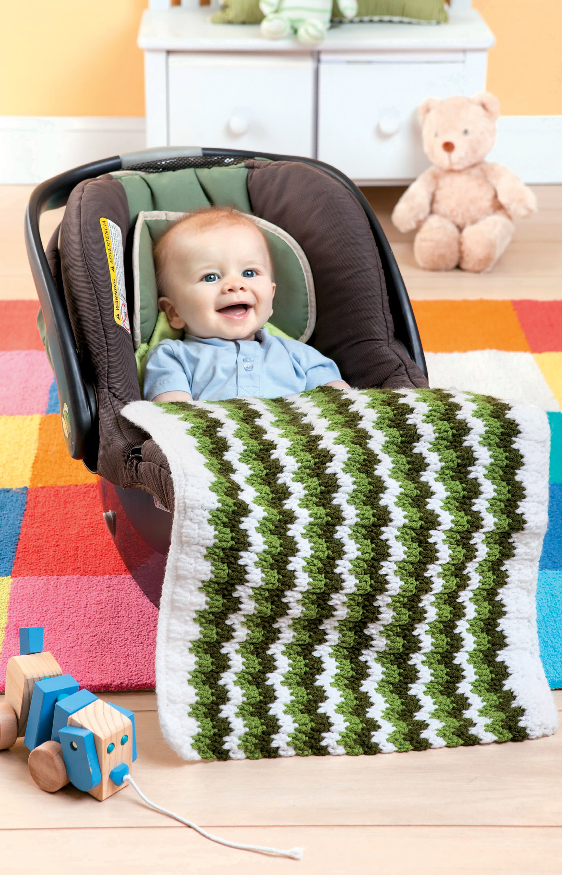A smiling baby sits in a car seat with a green and white zigzag-patterned blanket; Crochet Family Favorites by Leisure Arts, like colorful toys and a teddy bear, are on the rug in the background.