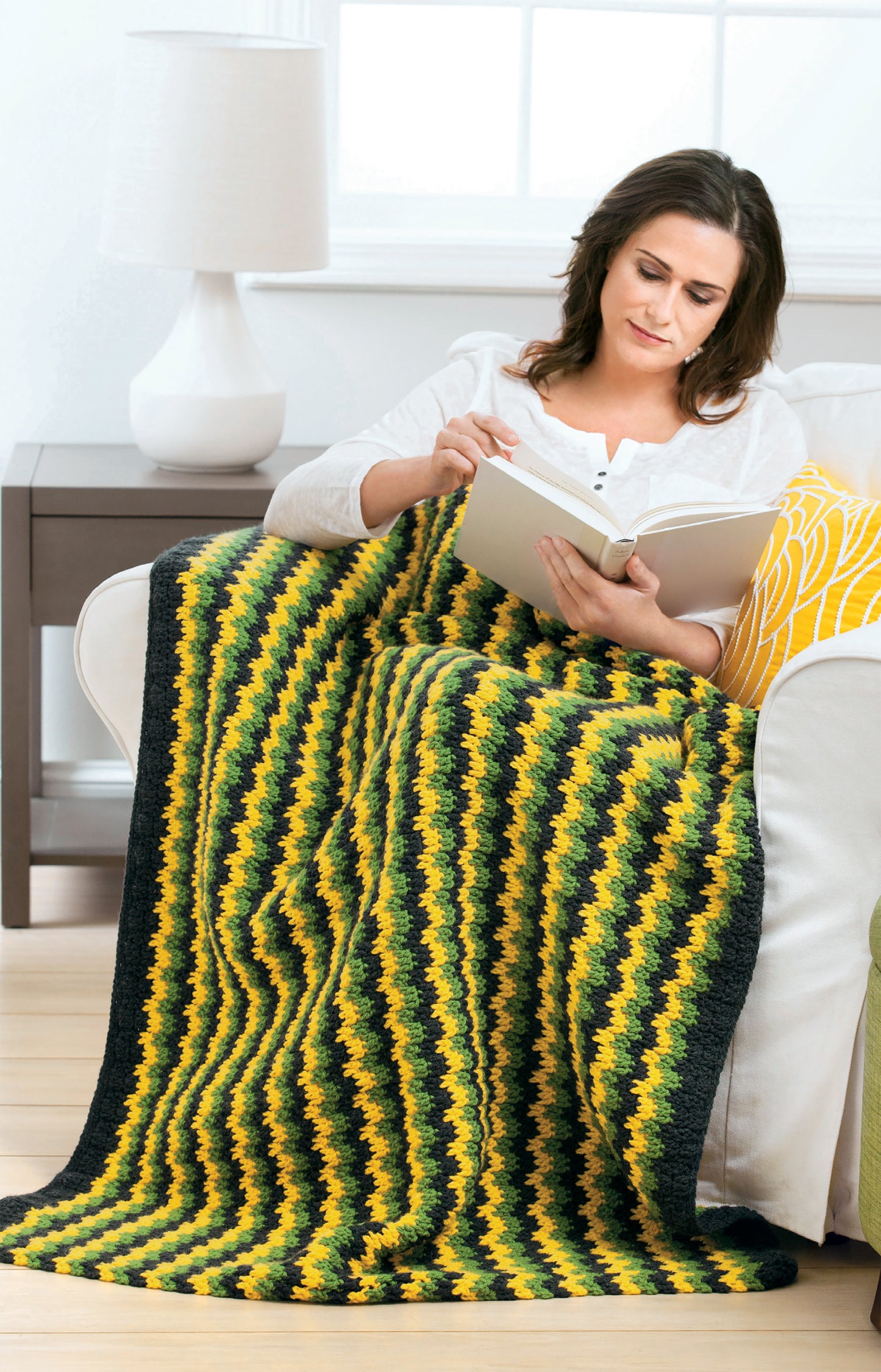 A woman relaxes on a white couch, reading with the Crochet Family Favorites blanket by Leisure Arts draped over her legs. Cozy slippers peek out as she sits beside a lamp and a yellow-patterned pillow.