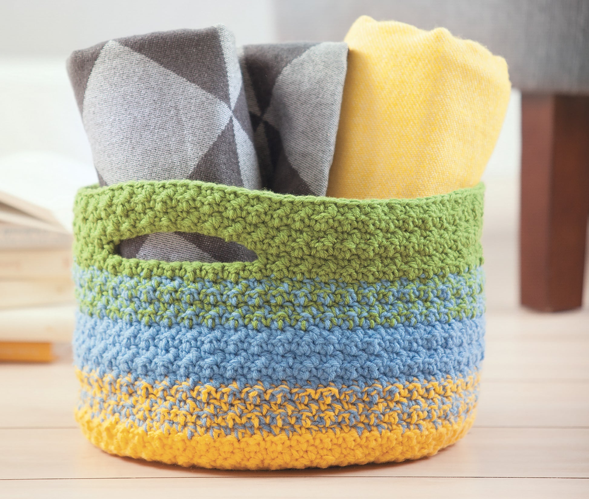 A colorful crocheted basket sits on a wooden floor, filled with scarves and blankets made using the Crochet Family Favorites Digital Download by Leisure Arts.
