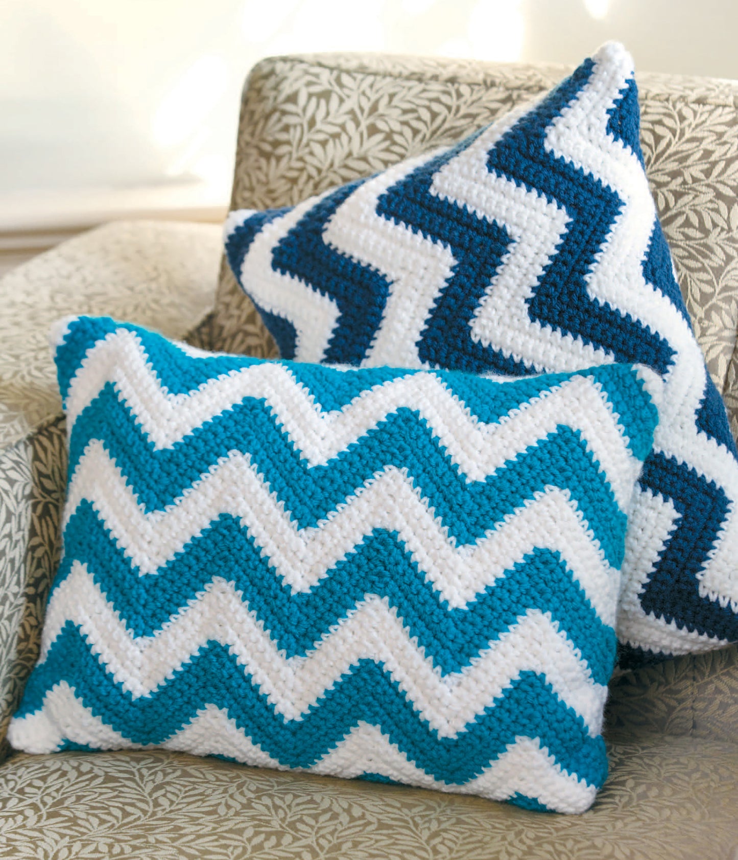 Two Leisure Arts Pillow Fun pillows featuring zigzag patterns in blue, white, and teal rest on a leafy beige couch—one upright, one leaning—adding playful, cozy accents to your home.