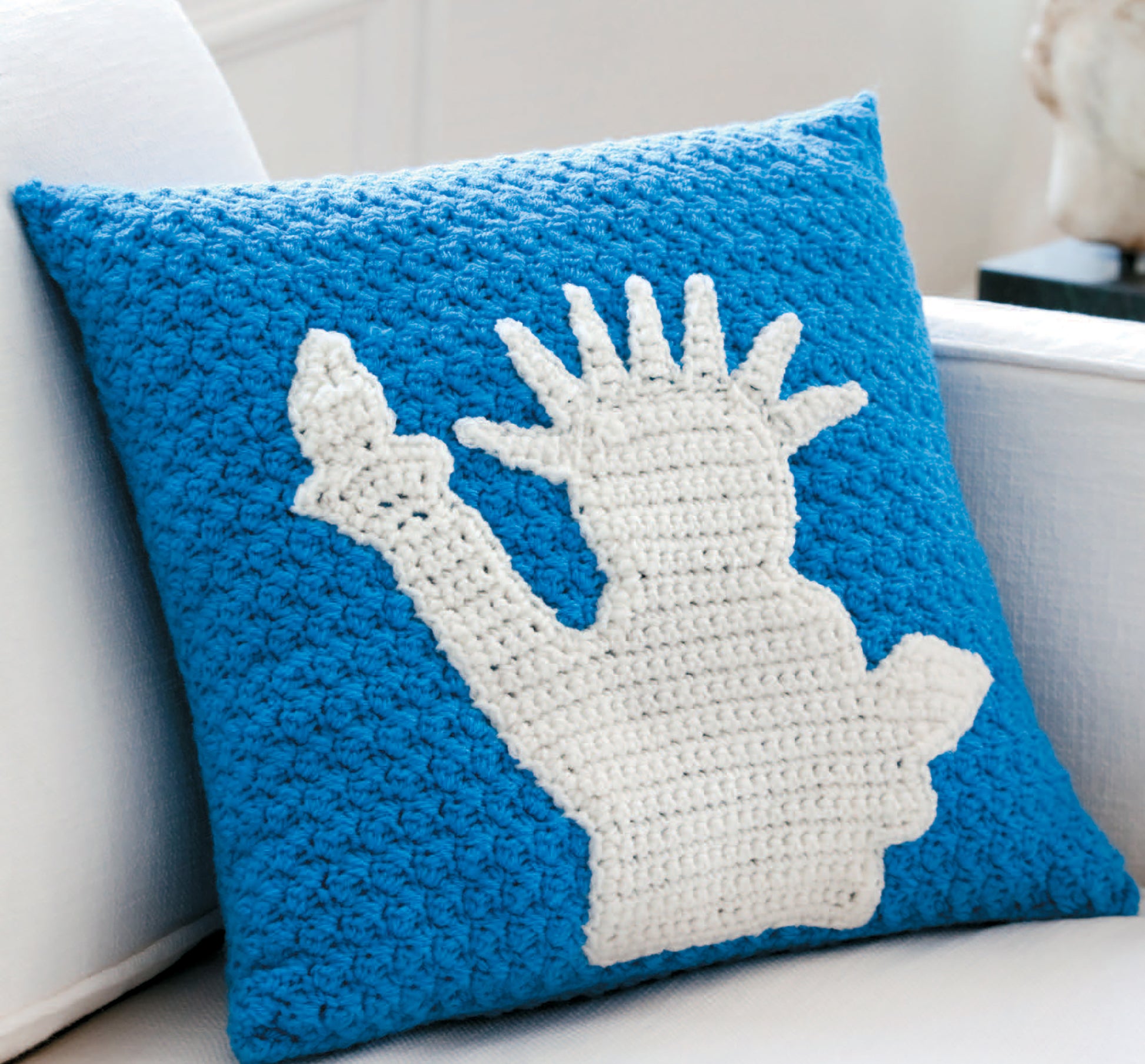 The Pillow Fun—Playful Accents for Your Home! from Leisure Arts is a blue crocheted pillow with a white Statue of Liberty silhouette, adding whimsy and charm to any space, especially when placed on a white couch.