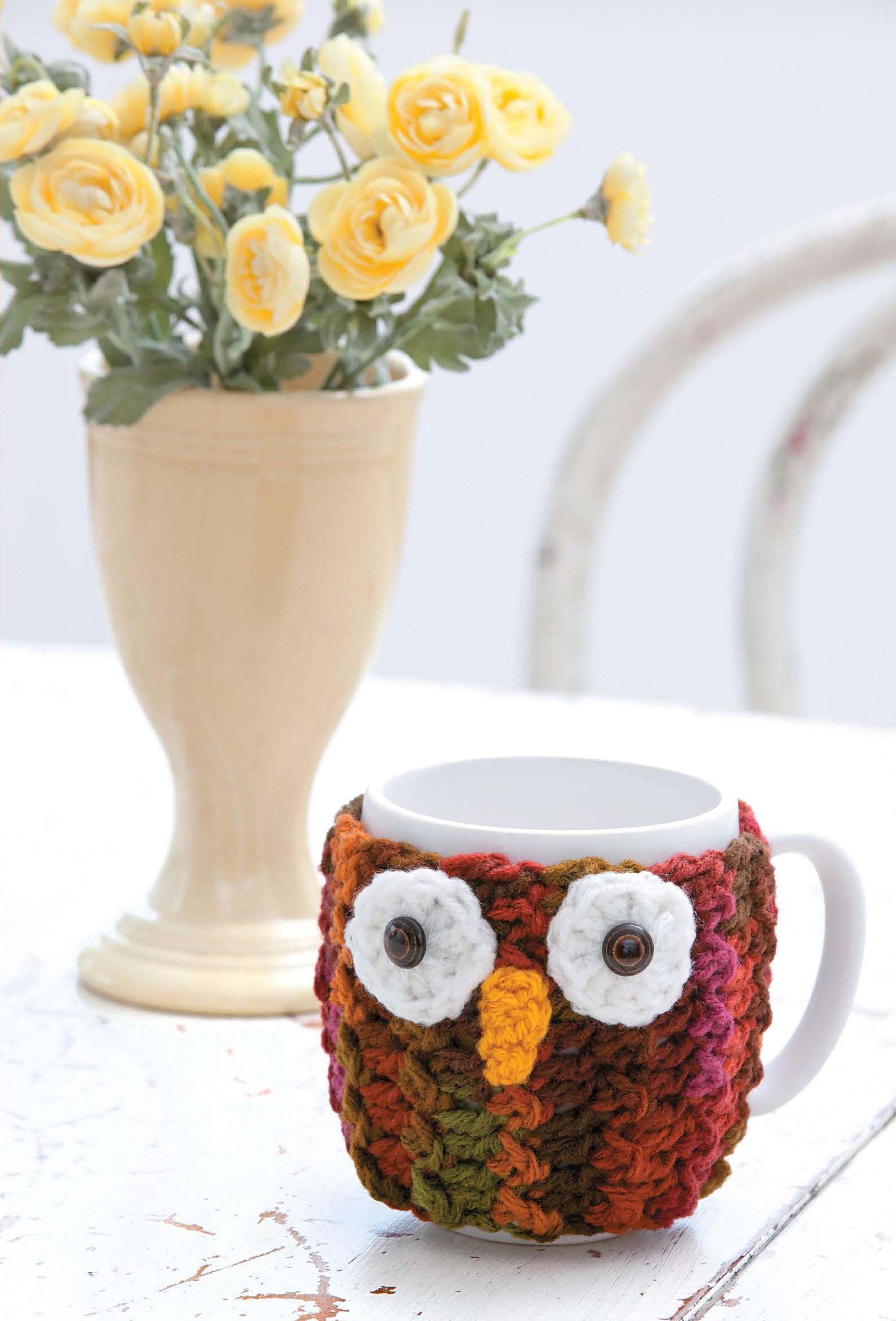 A Leisure Arts Mug Hugs & Coaster Set, featuring a colorful crocheted owl cozy with big white eyes and a yellow beak, sits on a white table beside snowflake coasters and a vase of yellow flowers.