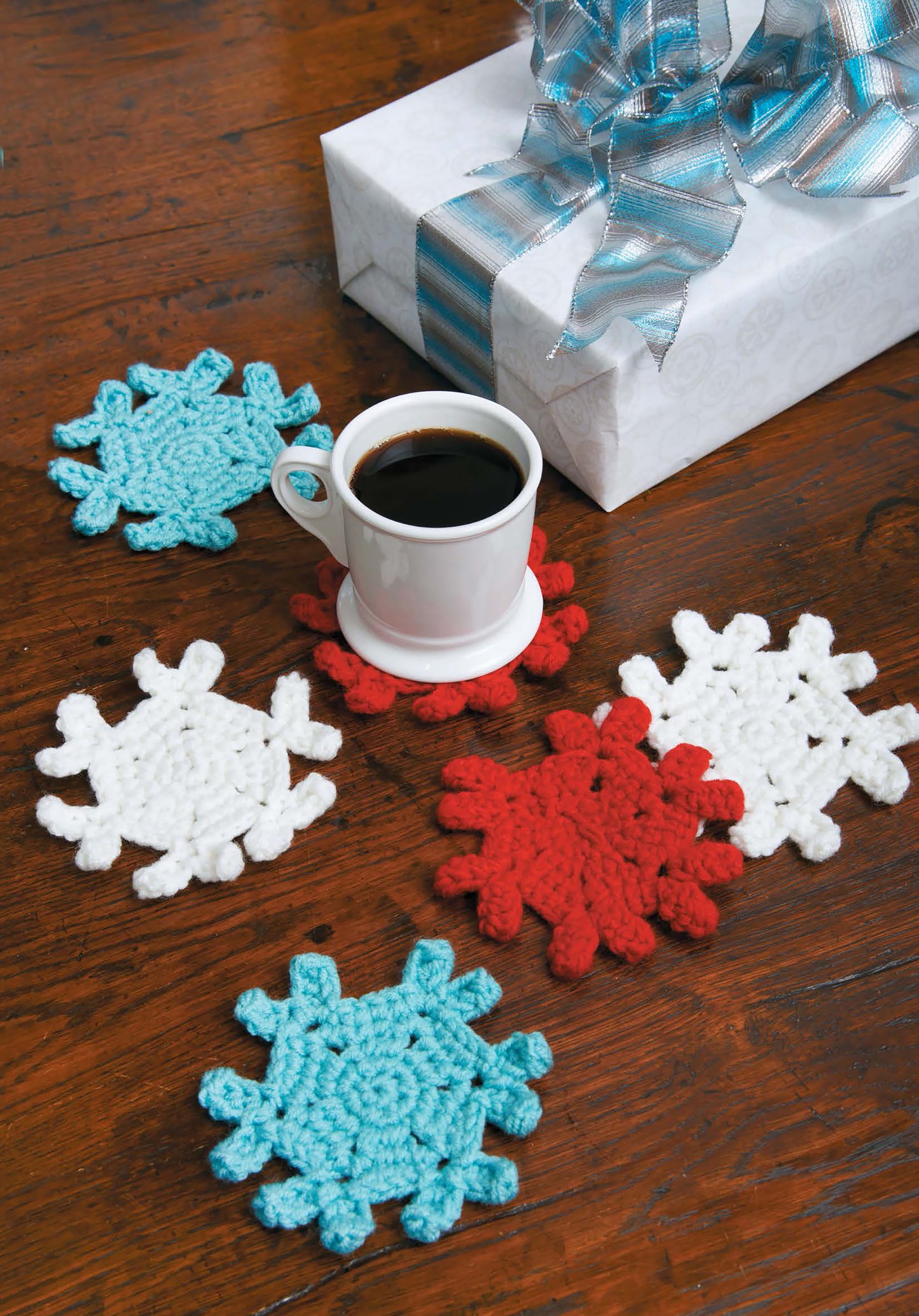 A cup of coffee rests on a red snowflake coaster from the Leisure Arts Mug Hugs & Coaster Sets, surrounded by festive blue, red, and white crocheted coasters on a wooden table. A wrapped gift with blue and silver ribbon sits nearby.