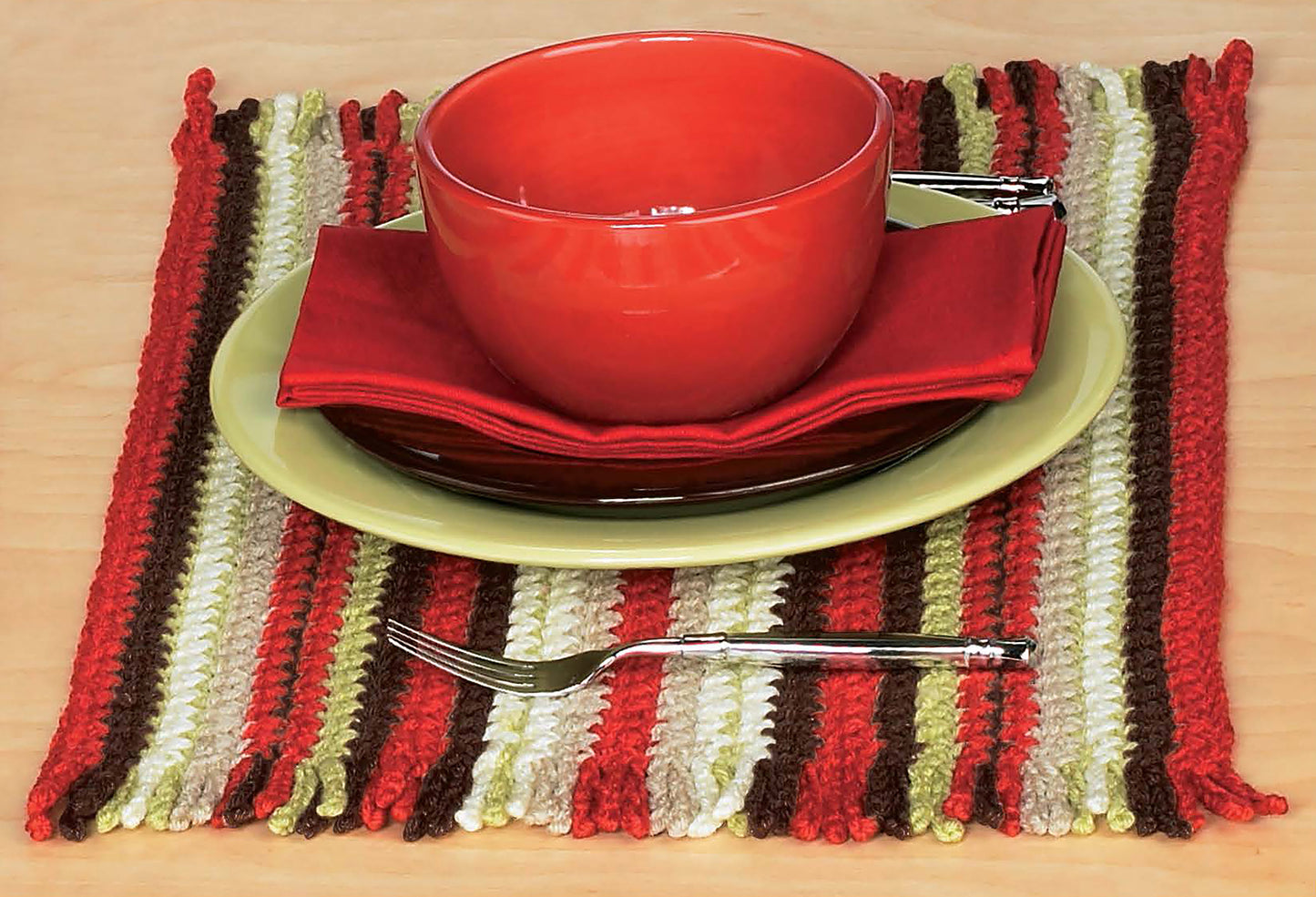 A Mug Hugs & Coaster Set by Leisure Arts sits on a green plate and red napkin atop a striped, crochet-inspired placemat in red, brown, green, and white. A fork rests in front on the wooden table.