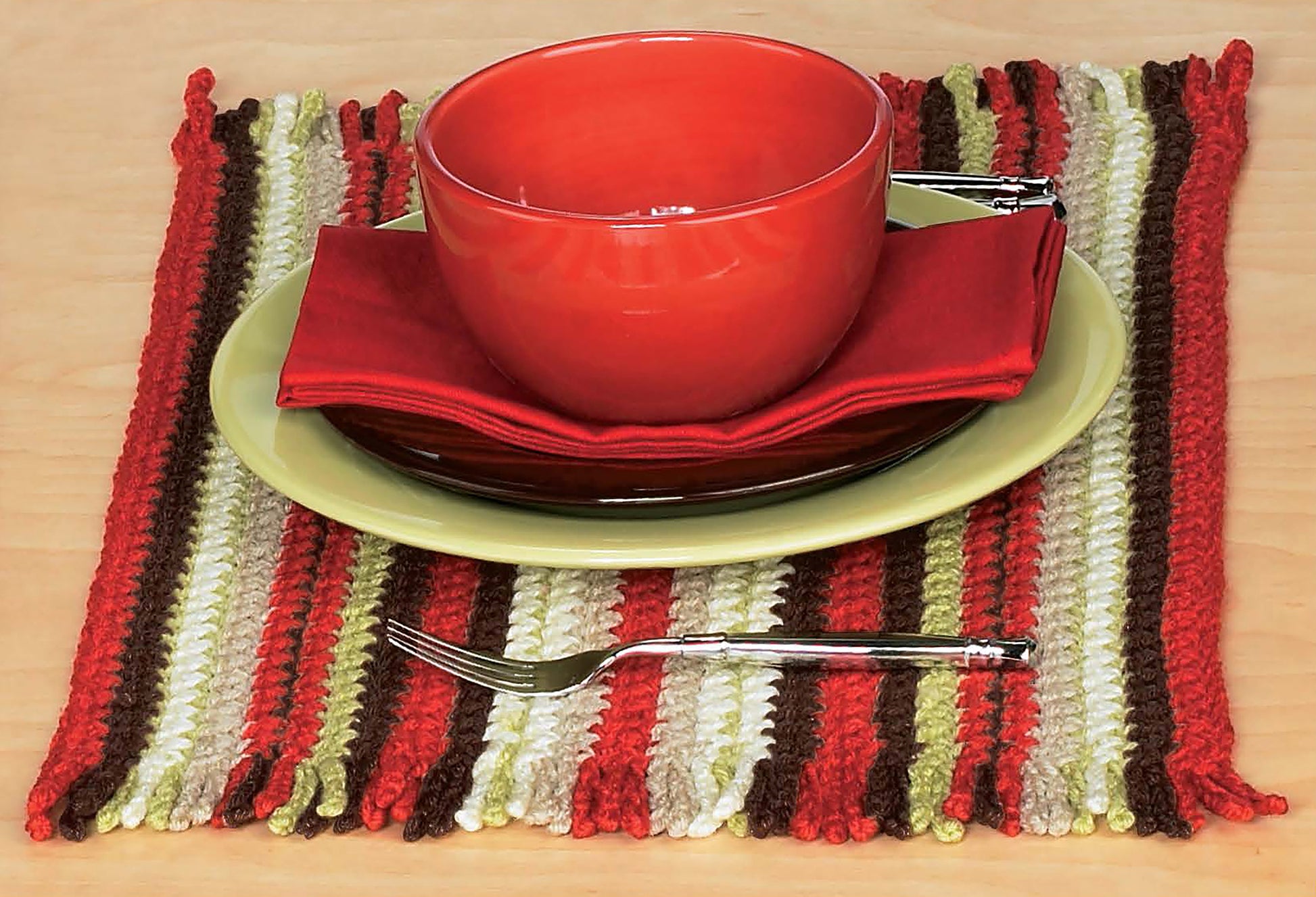 A Mug Hugs & Coaster Set by Leisure Arts sits on a green plate and red napkin atop a striped, crochet-inspired placemat in red, brown, green, and white. A fork rests in front on the wooden table.