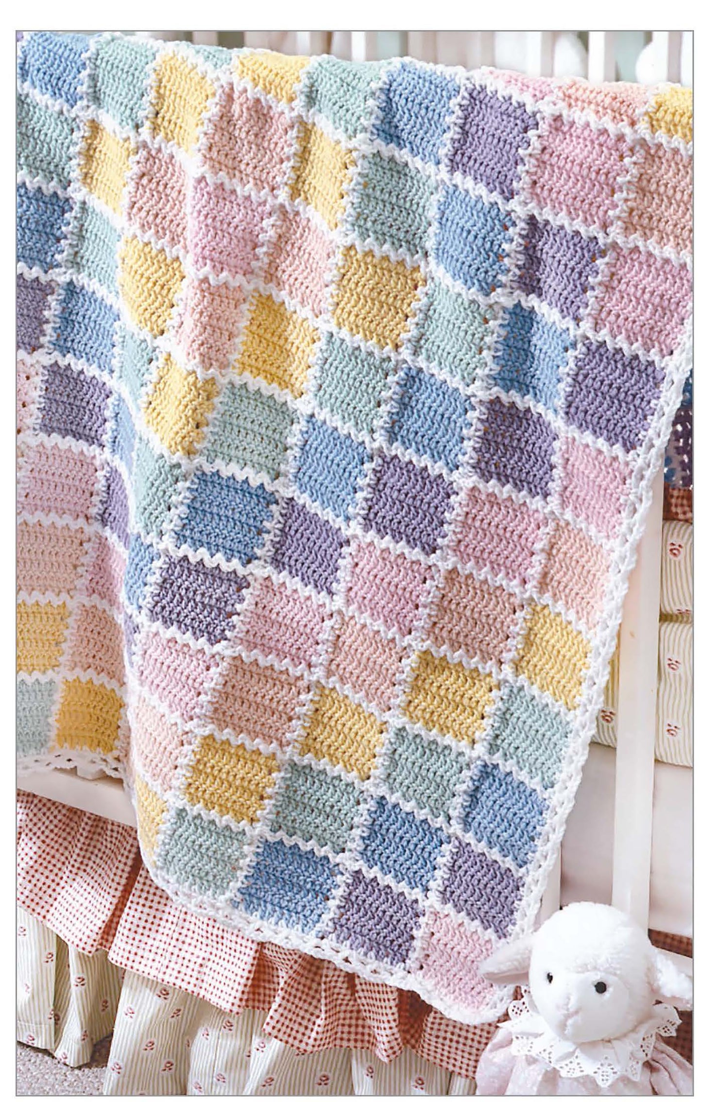 A pastel, checkerboard-patterned baby blanket—crocheted with inspiration from "The Little Encyclopedia of Crocheted Baby Blankets" by Leisure Arts—draped over a white crib beside a plush white lamb toy.