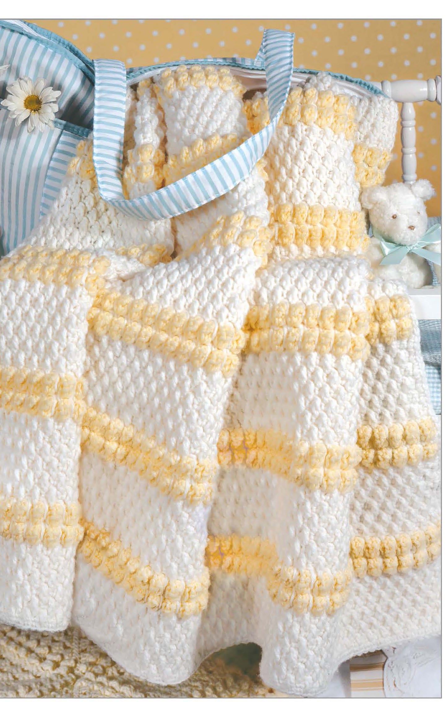 A soft white and yellow baby blanket, inspired by patterns from The Little Encyclopedia of Crocheted Baby Blankets by Leisure Arts, is draped over a chair beside a blue-striped bag, teddy bear, polka dot wallpaper, and daisies.