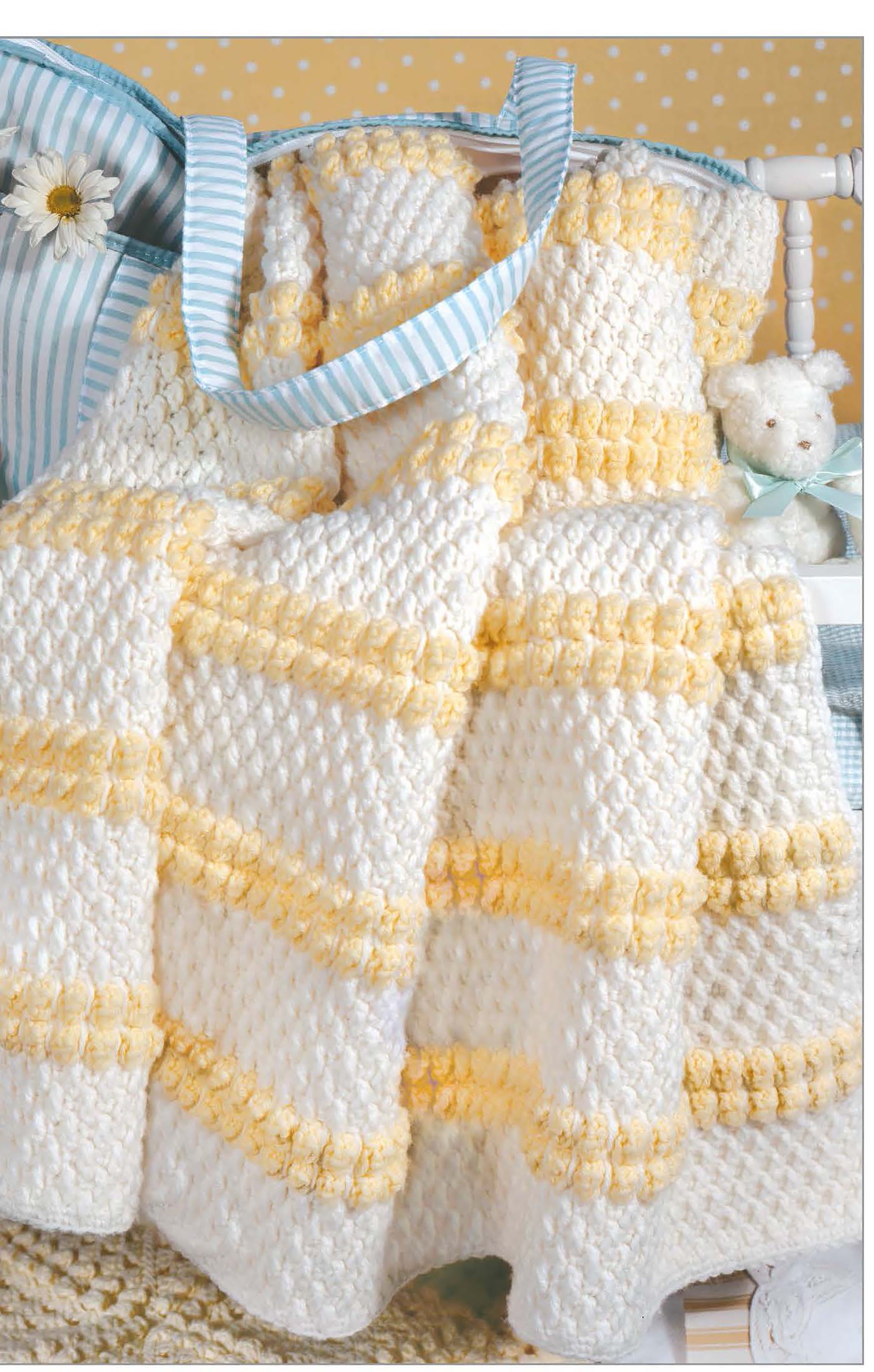 A soft white and yellow baby blanket, inspired by patterns from The Little Encyclopedia of Crocheted Baby Blankets by Leisure Arts, is draped over a chair beside a blue-striped bag, teddy bear, polka dot wallpaper, and daisies.