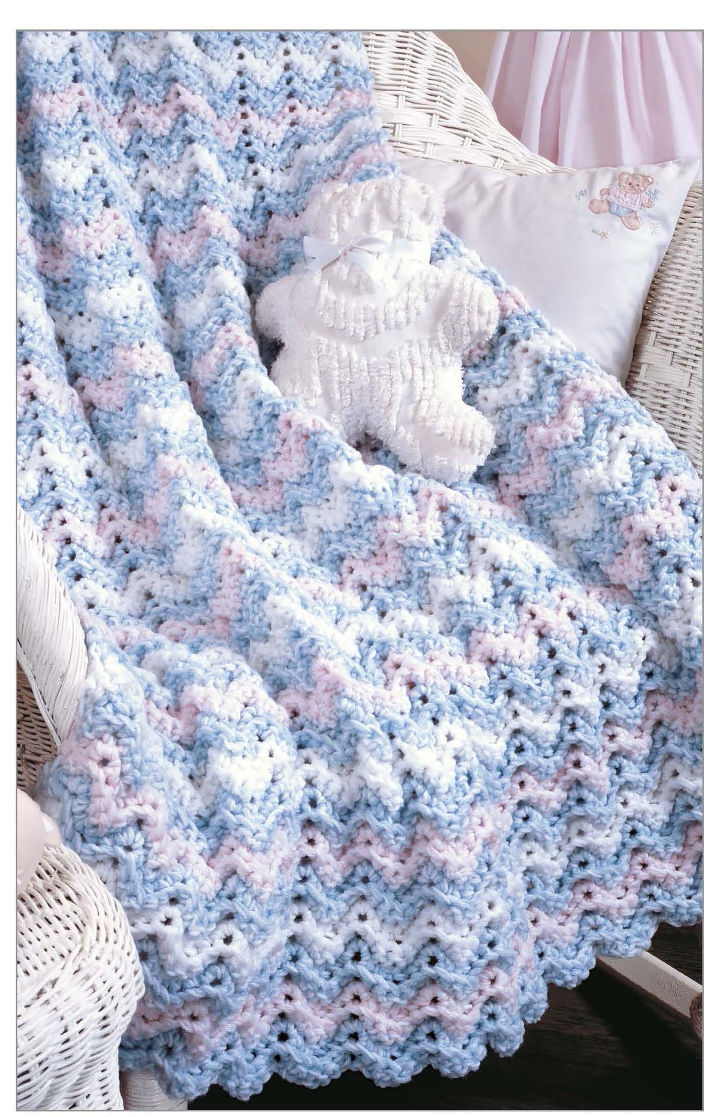 The Little Encyclopedia of Crocheted Baby Blankets by Leisure Arts is displayed with a pastel blue, pink, and white zigzag-patterned blanket draped over a white wicker chair, topped with a textured white teddy bear.