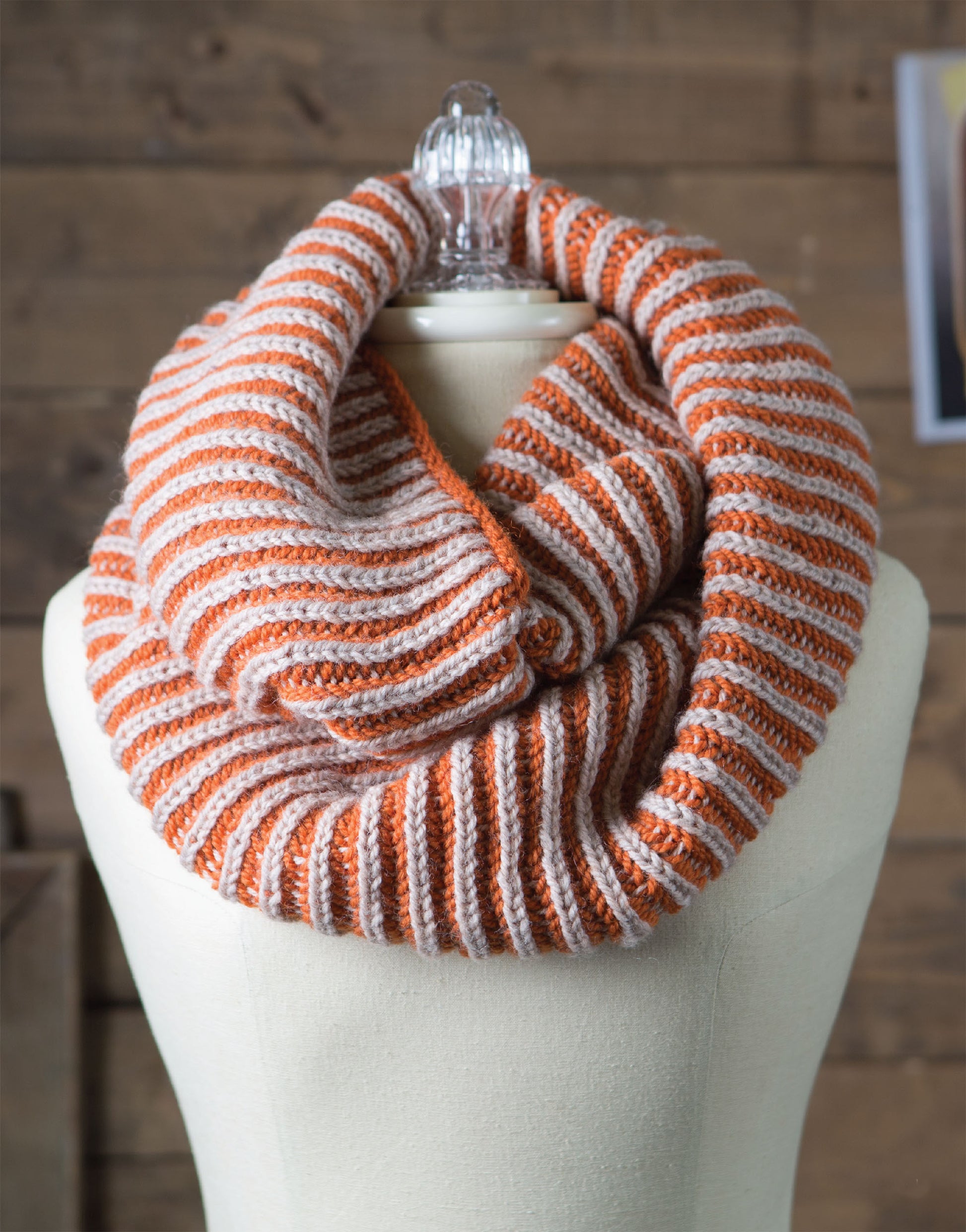A cream mannequin showcases Leisure Arts' Brioche Knitting infinity scarf in orange and beige stripes, displayed against a wooden background.
