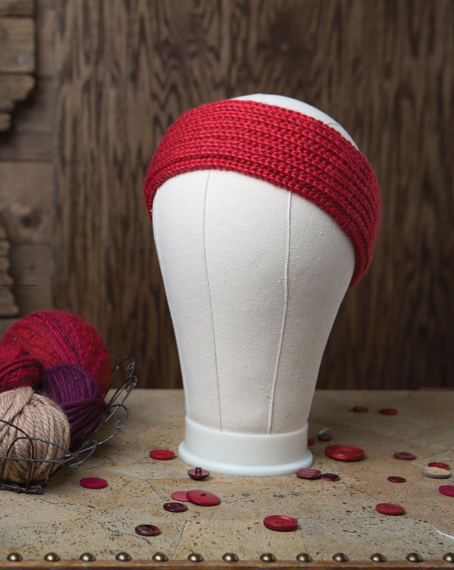 A white mannequin head models a thick, brioche knit red headband made with Leisure Arts' Brioche Knitting. Red buttons and yarn in red and beige sit on a textured surface before a rustic wooden backdrop.