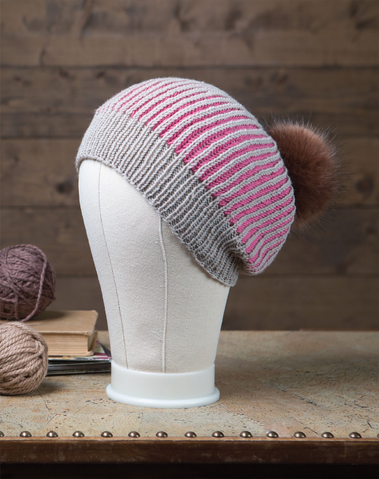 The Brioche Knitting hat by Leisure Arts features gray and pink stripes in a brioche style with a large brown pom-pom, displayed on a white mannequin head. Yarn balls and books for beginner knitters sit nearby on the wooden table.