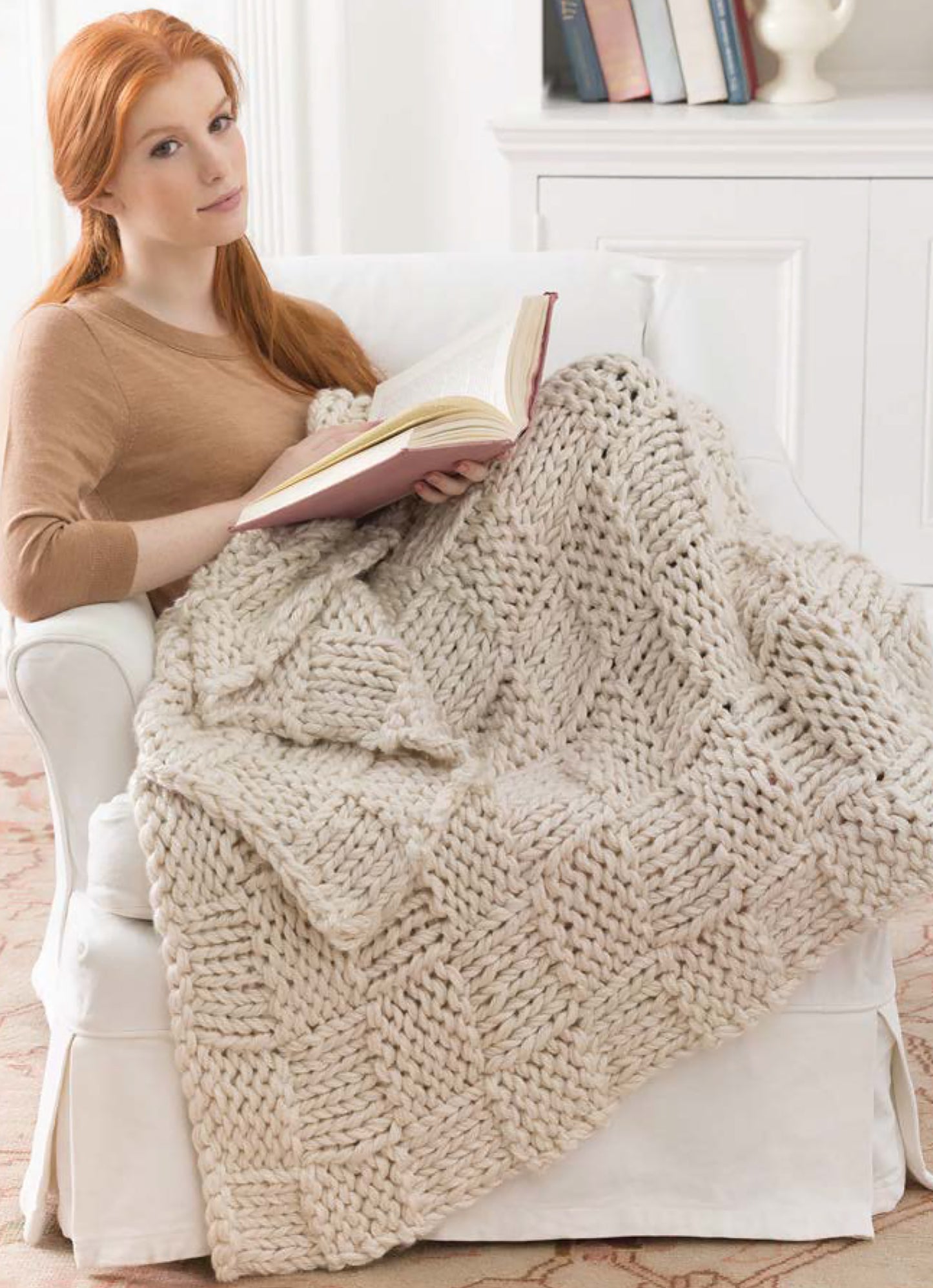 A woman with long red hair sits on a white armchair draped in a chunky beige knit blanket, reading "Project Knitwell Presents The Comfort of Knitting" by Leisure Arts. Books line the shelves in the background.