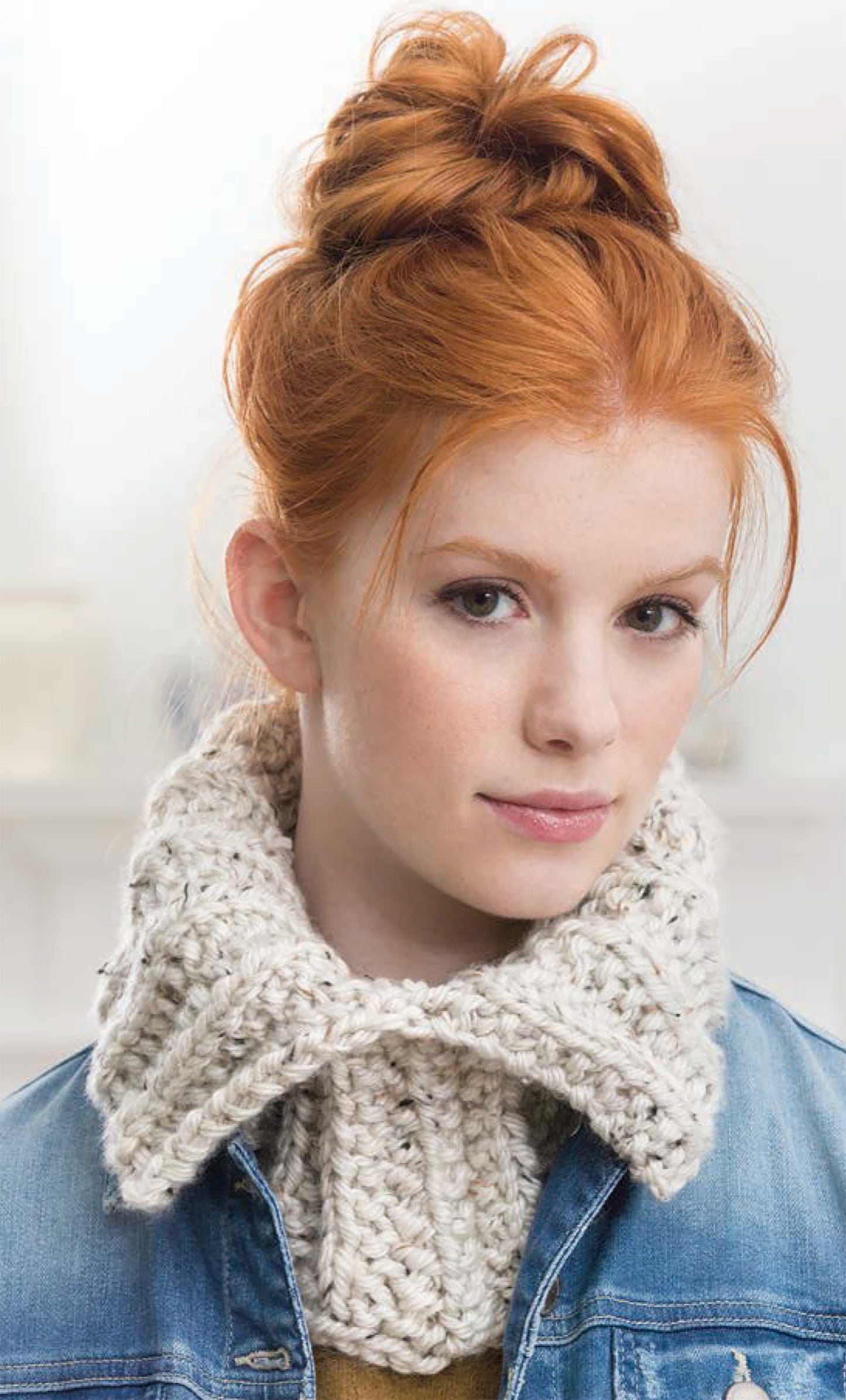 A young woman with fair skin and red hair in a messy bun wears a blue denim jacket and a chunky, cream knitted scarf—perfect inspiration for easy projects from "Project Knitwell Presents The Comfort of Knitting" by Leisure Arts. The background is softly blurred.