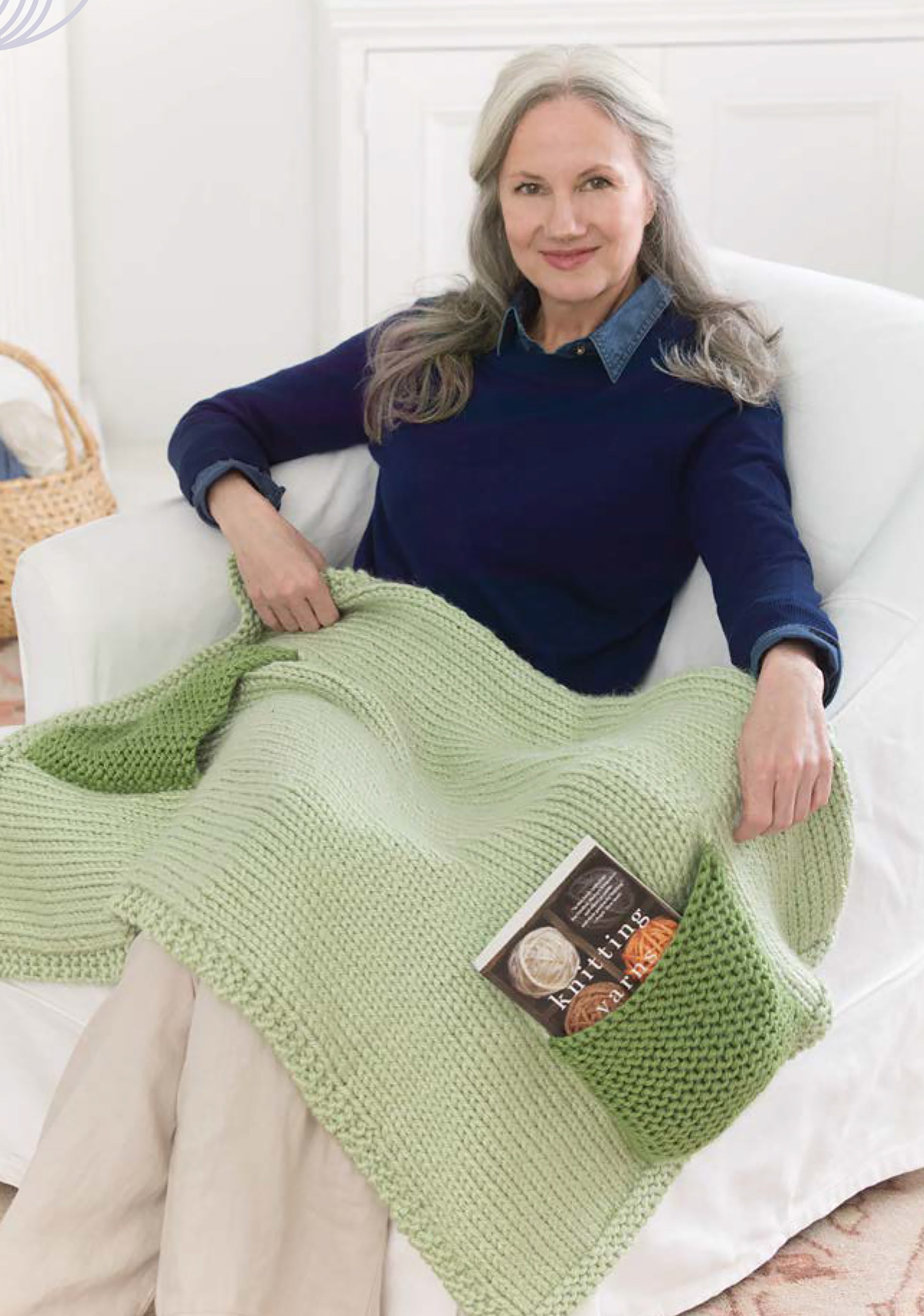 A woman with long gray hair, in a navy sweater and beige pants, smiles on a white chair. A green knitted Leisure Arts “Project Knitwell Presents The Comfort of Knitting” blanket with a book in its pocket is draped over her lap—perfect for easy projects.