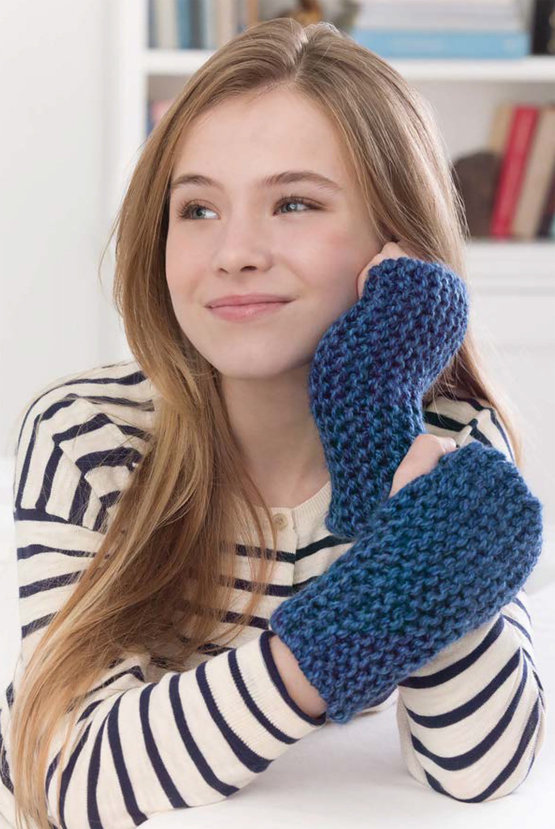 A young girl with long blonde hair, wearing a black-and-white striped shirt and blue fingerless gloves from "Project Knitwell Presents The Comfort of Knitting" by Leisure Arts, rests her chin on her arms and smiles thoughtfully. Bookshelves are in the background.