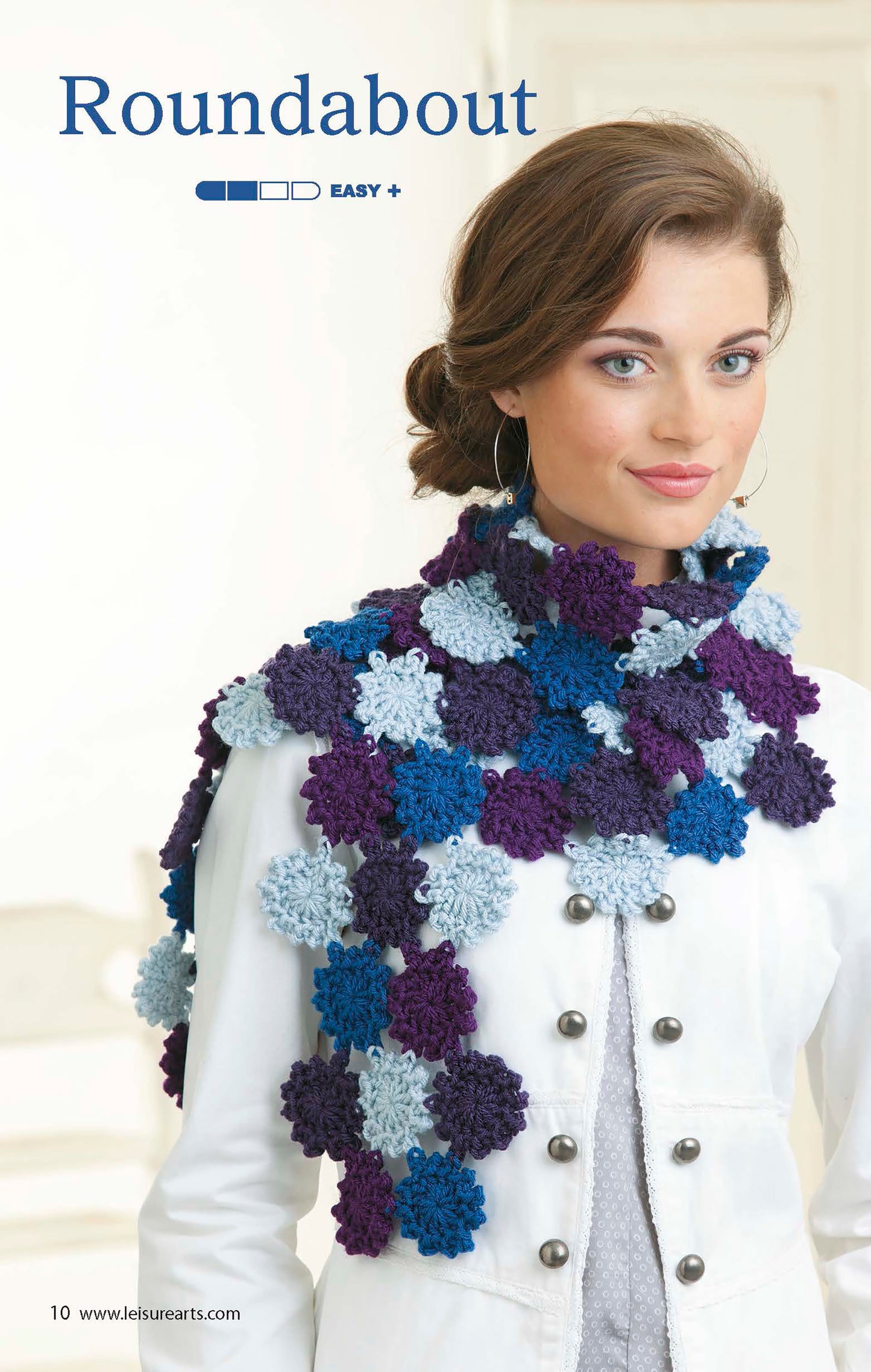 A woman in a white jacket models a blue and purple, flower-clustered scarf from The Little Encyclopedia of Crocheted Scarves digital download by Leisure Arts—an easy, creative choice for anyone seeking beginner crochet scarf patterns.