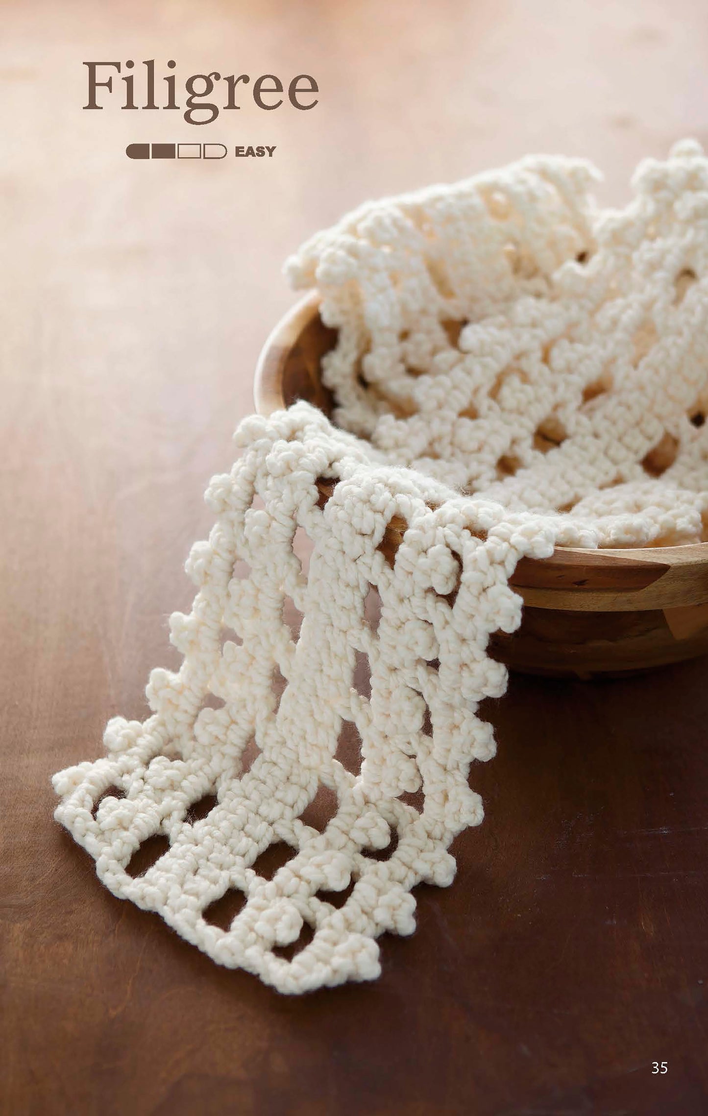 A cream-colored, crocheted filigree pattern drapes over a wooden bowl on a wooden surface. At the top are the words "The Little Encyclopedia of Crocheted Scarves" by Leisure Arts and an easy skill level indicator.