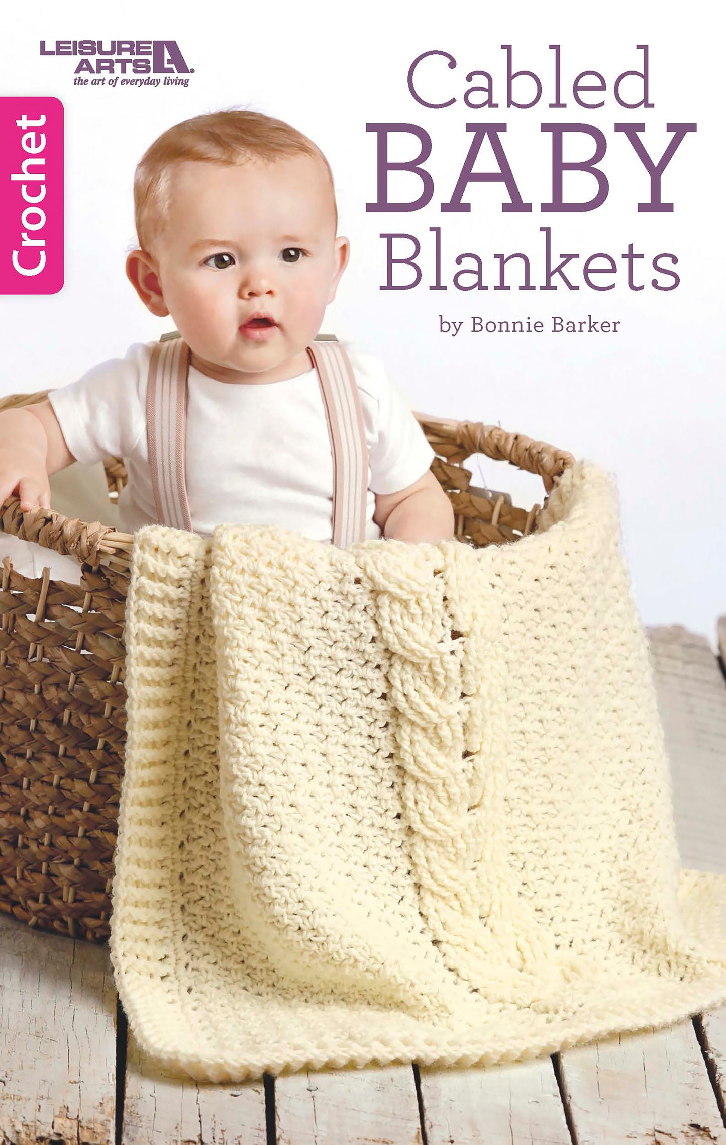 A baby sits in a woven basket with a cream crocheted blanket. The book "Cabled Baby Blankets" by Leisure Arts offers intermediate crochet baby afghan patterns using worsted weight yarn.