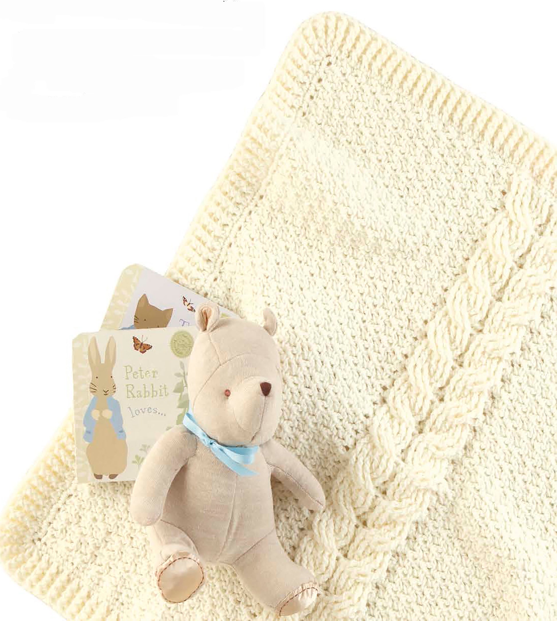 The Leisure Arts Cabled Baby Blankets in cream, paired with a beige teddy bear with a blue ribbon and two Peter Rabbit-themed board books, are arranged together on a white background.