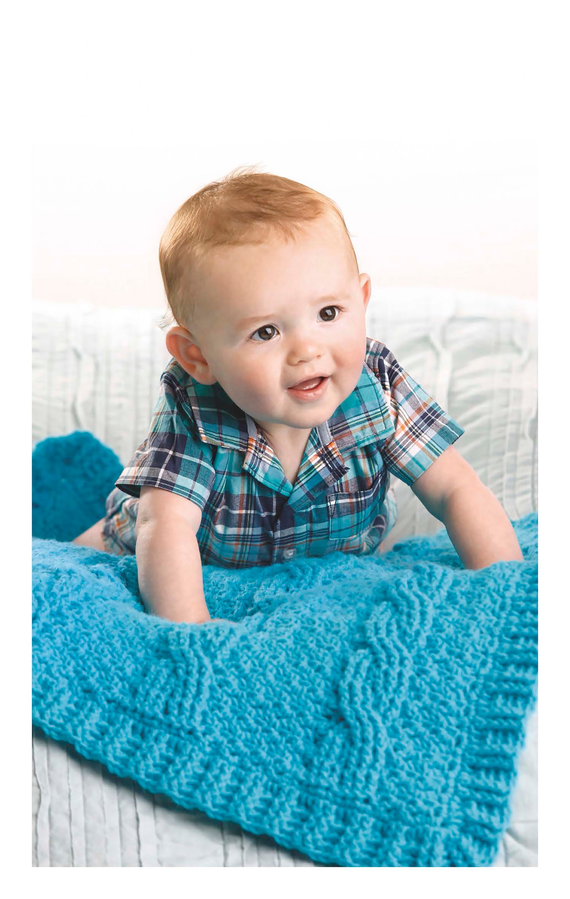 A smiling baby with light hair lies on a vibrant blue Cabled Baby Blanket from Leisure Arts, made with worsted weight yarn—an ideal choice for those seeking an intermediate crochet project.