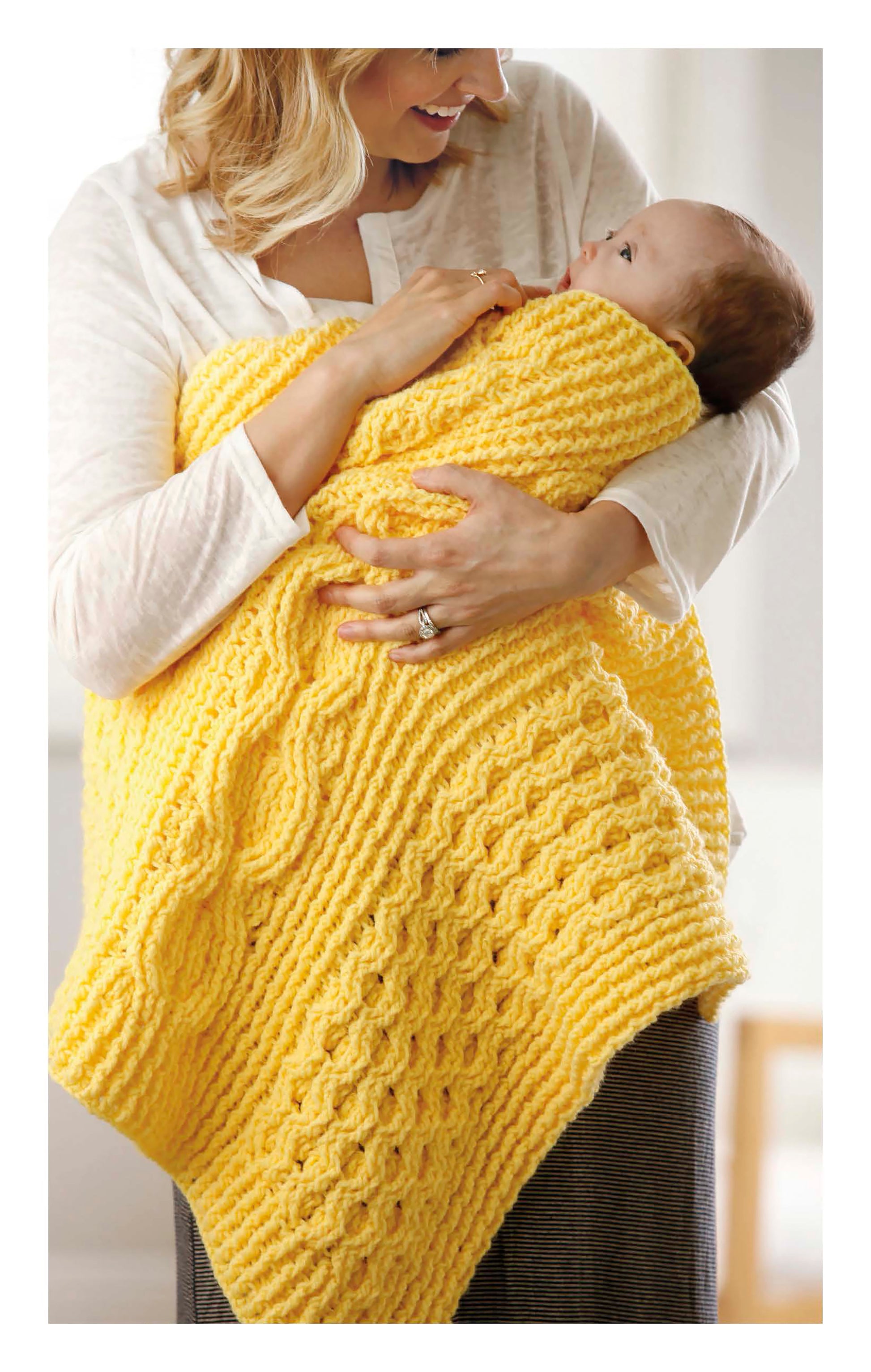 A woman smiles at a baby wrapped in a bright yellow cabled blanket made from worsted weight yarn. She holds the child close, highlighting Cabled Baby Blankets by Leisure Arts, perfect inspiration for intermediate crochet projects.