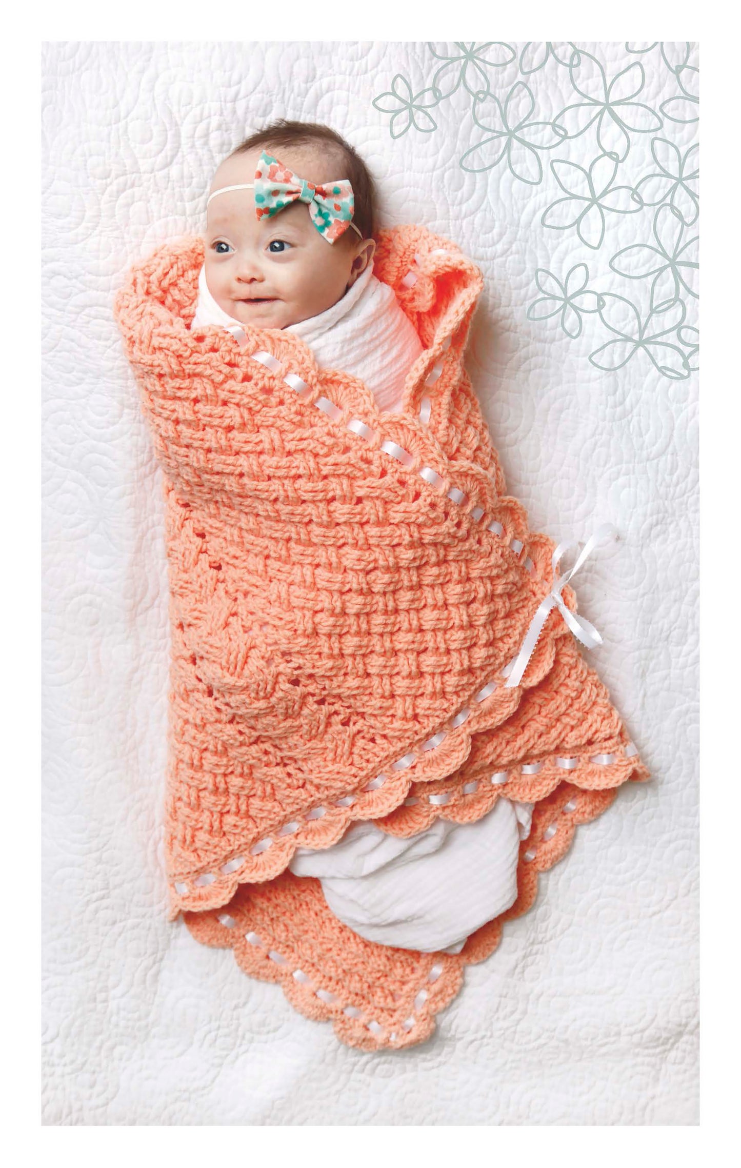 A baby wrapped in a peach Cabled Baby Blanket from Leisure Arts lies on a white quilt, wearing a floral headband and smiling up. Decorative floral patterns appear in the top right corner.