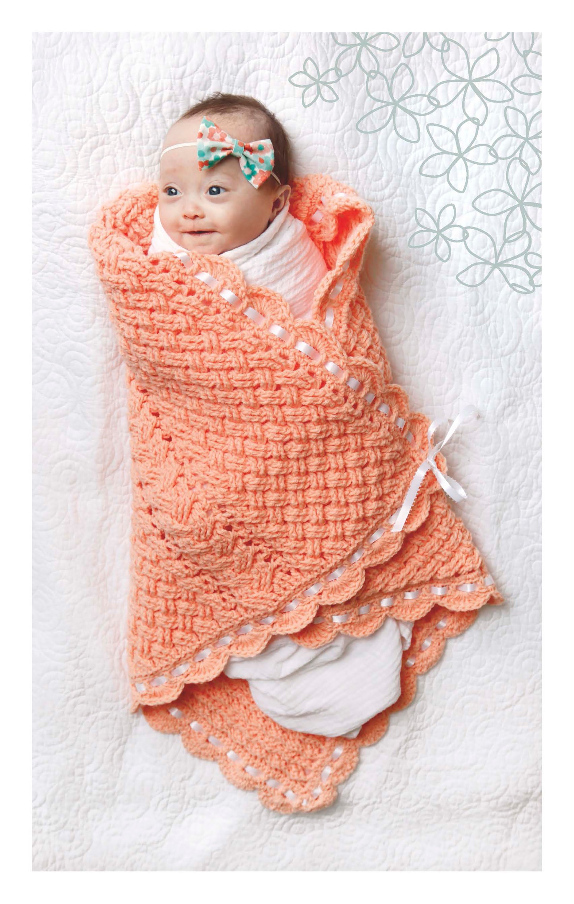 A baby wrapped in a peach Cabled Baby Blanket from Leisure Arts lies on a white quilt, wearing a floral headband and smiling up. Decorative floral patterns appear in the top right corner.