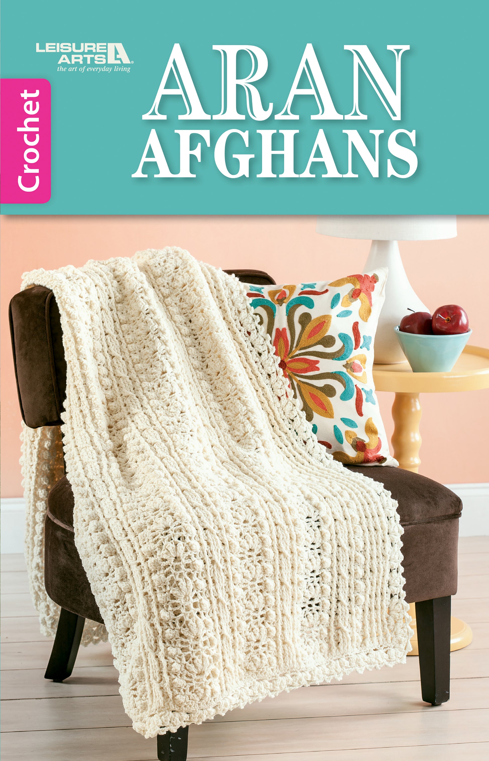 A cream Aran crochet afghan is draped over a brown chair with a floral pillow, next to a white table holding apples and a yellow cup. This cozy scene appears on the cover of "Aran Afghans" by Leisure Arts.