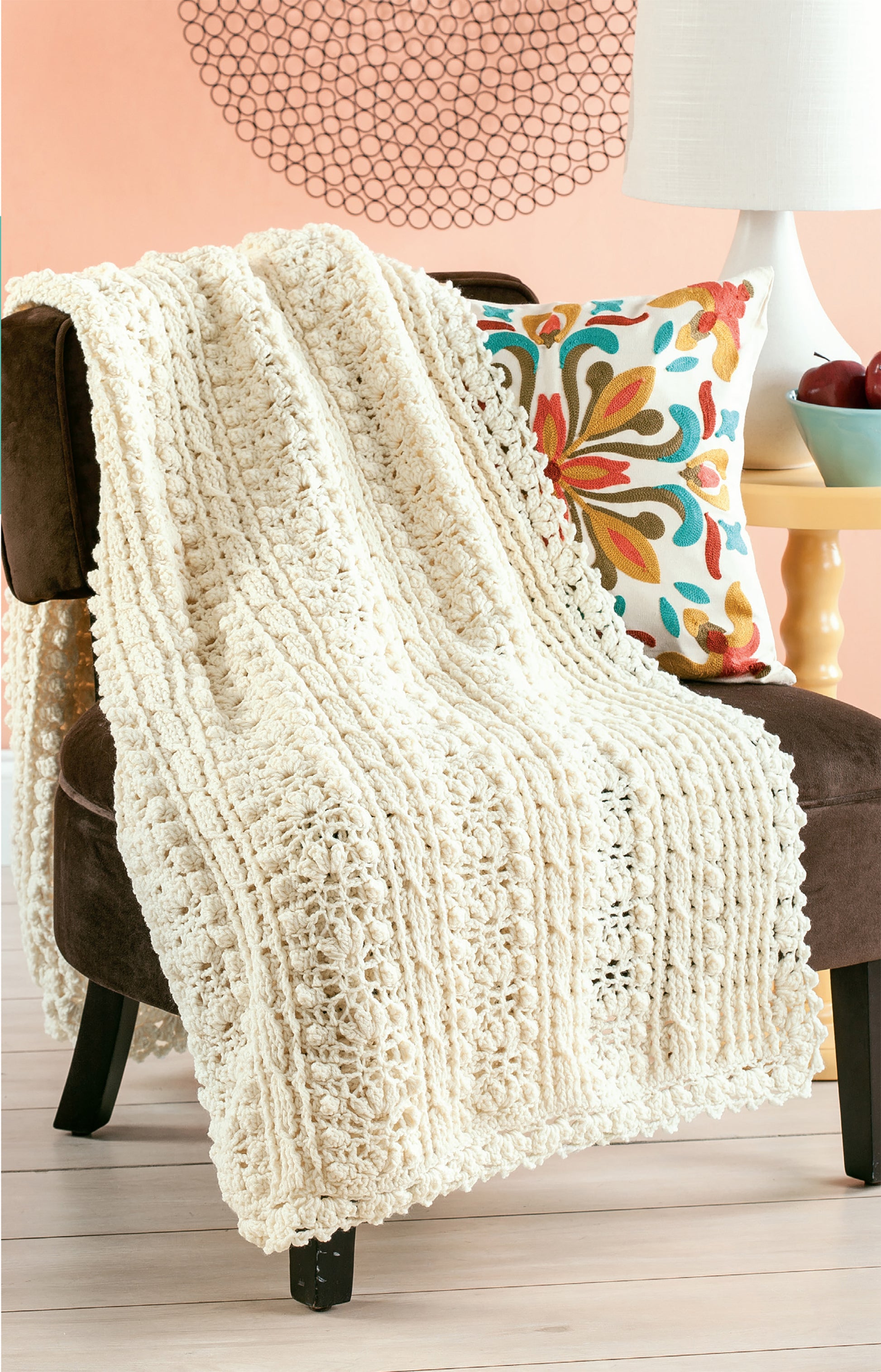 A cream Aran Afghans blanket by Leisure Arts is draped over a brown chair with a floral pillow, next to a yellow table and white lamp, set against a peach wall featuring a decorative circular pattern.