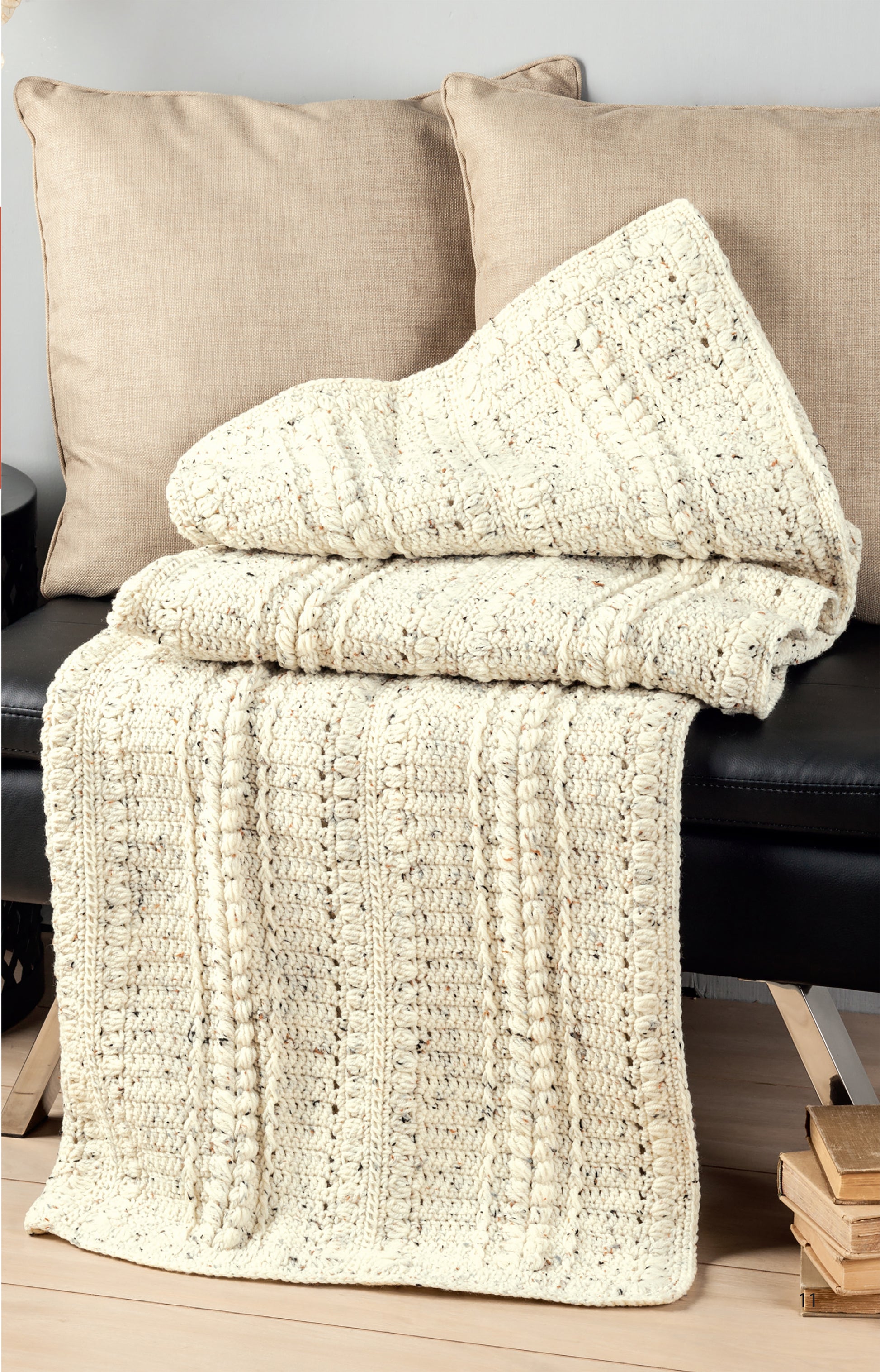 A cream-colored Aran Afghans crochet blanket by Leisure Arts is draped over a black bench with two beige pillows behind it, with wooden books stacked on the floor beside the bench.