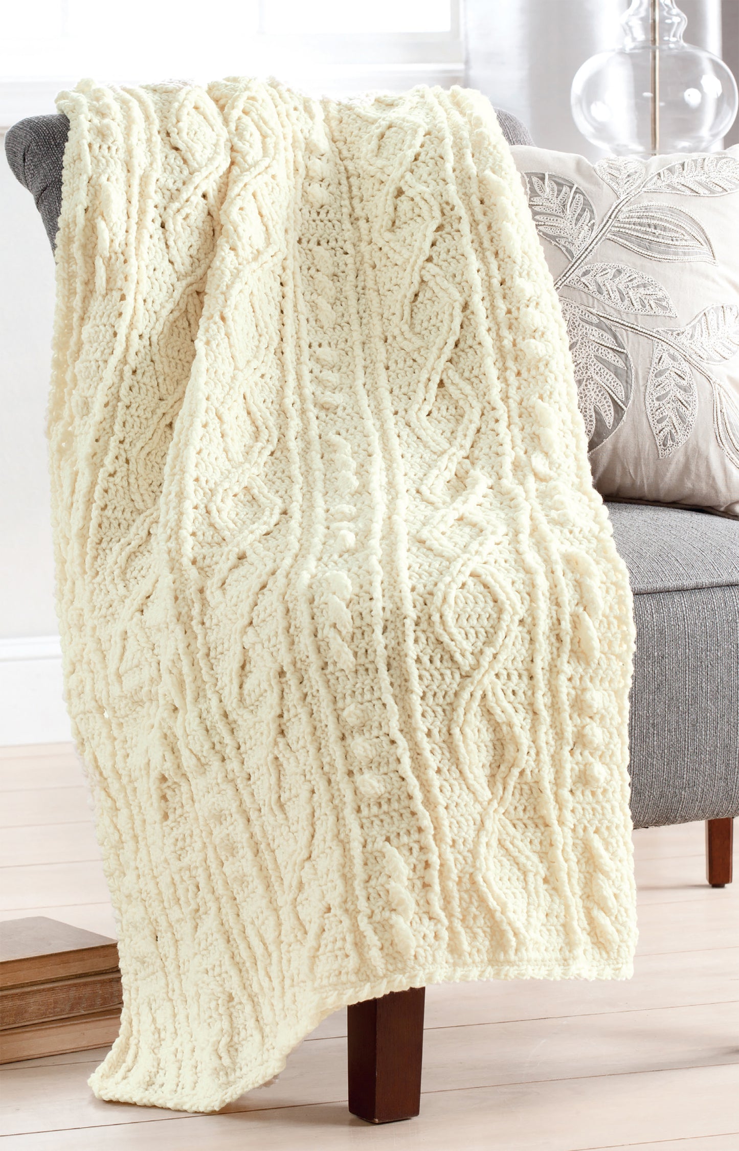 An Aran Afghans blanket by Leisure Arts, cream-colored with a fisherman knit, drapes over a gray upholstered chair beside a leaf-patterned pillow in a bright, cozy room with wooden floors.