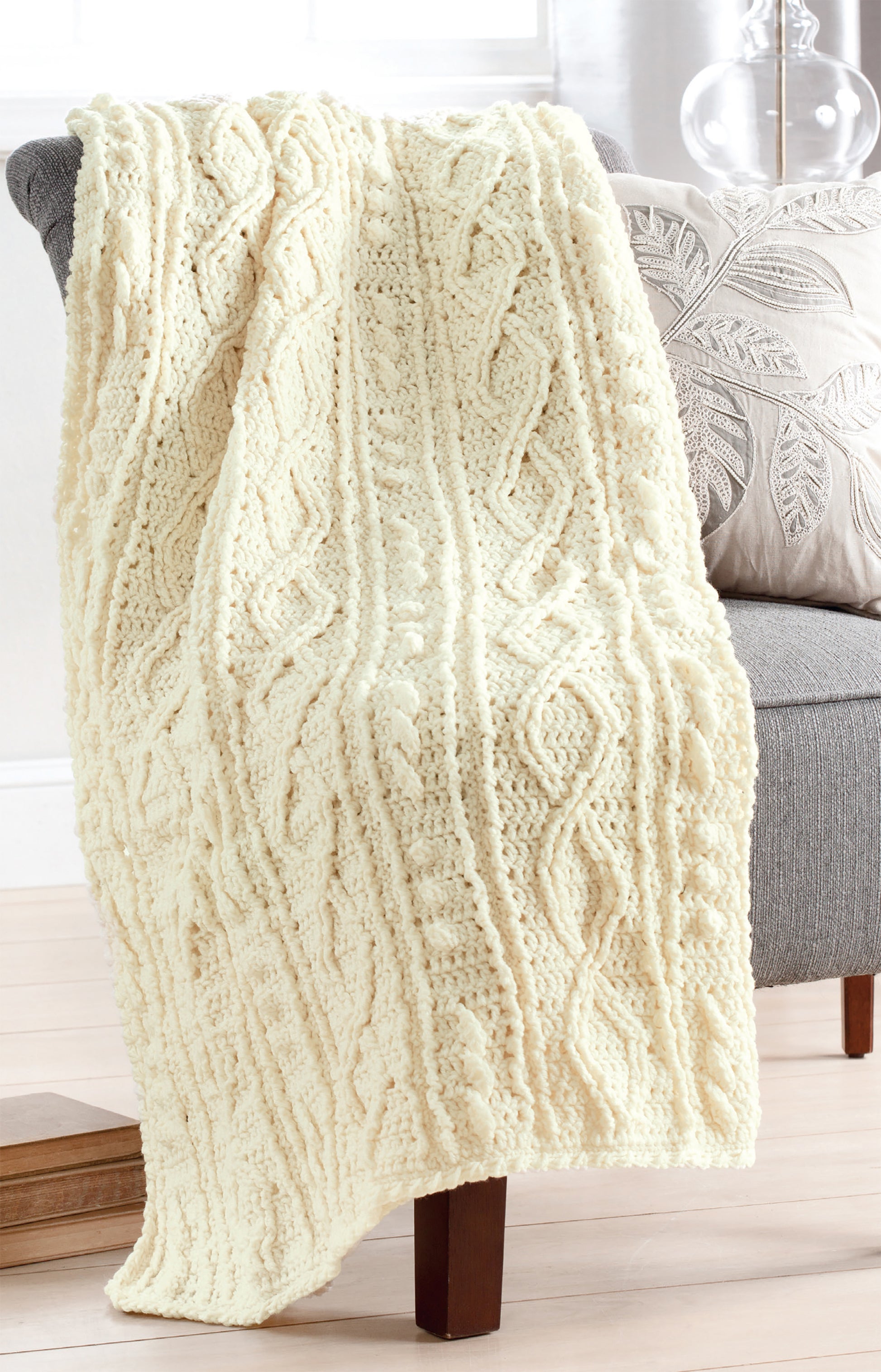 An Aran Afghans blanket by Leisure Arts, cream-colored with a fisherman knit, drapes over a gray upholstered chair beside a leaf-patterned pillow in a bright, cozy room with wooden floors.