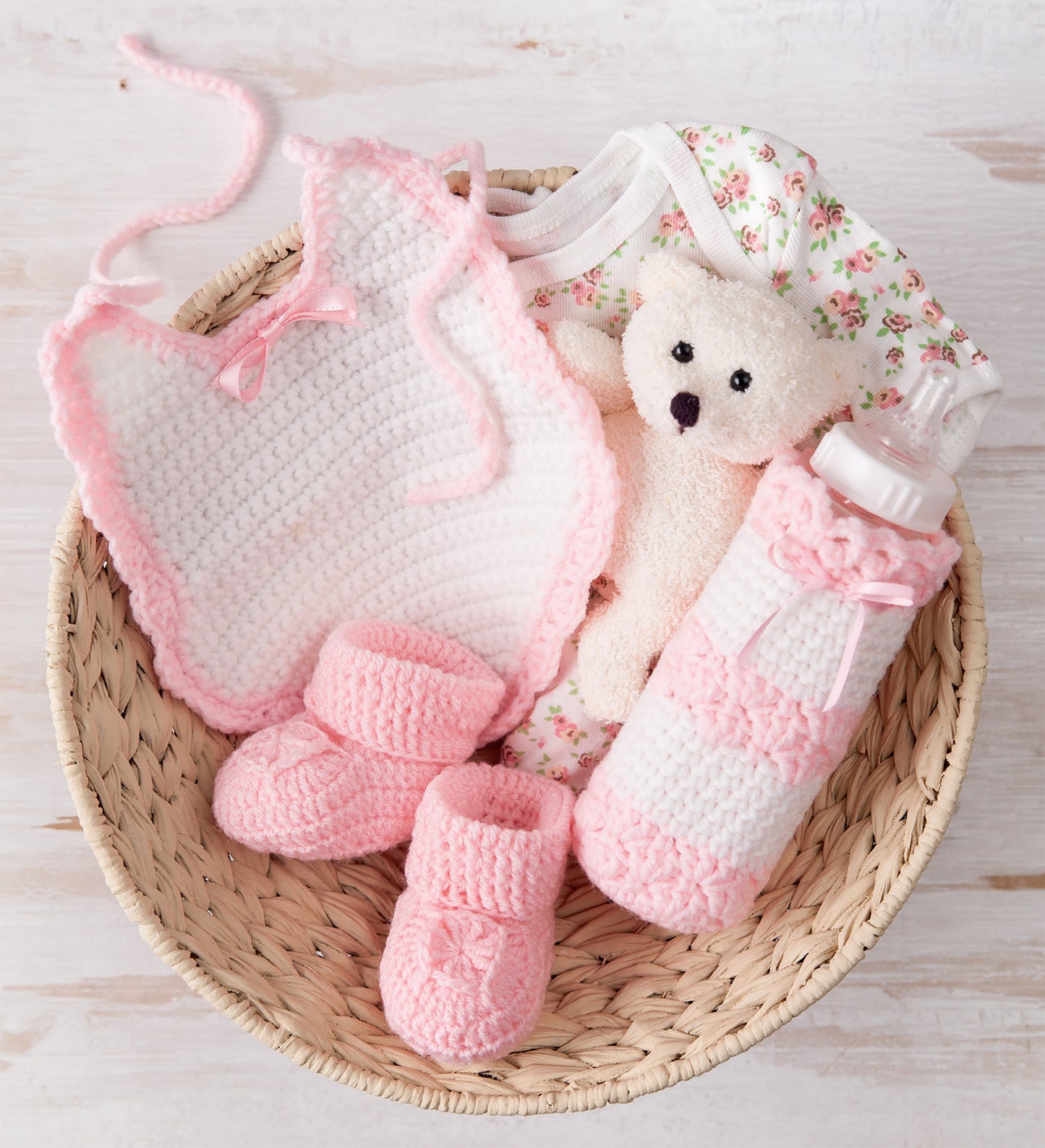 The Leisure Arts Bibs, Bottle Covers & Booties set features a woven basket with pink crocheted booties, a white and pink bib, floral onesie, small white teddy bear, and baby bottle adorned with a charming bottle cover.