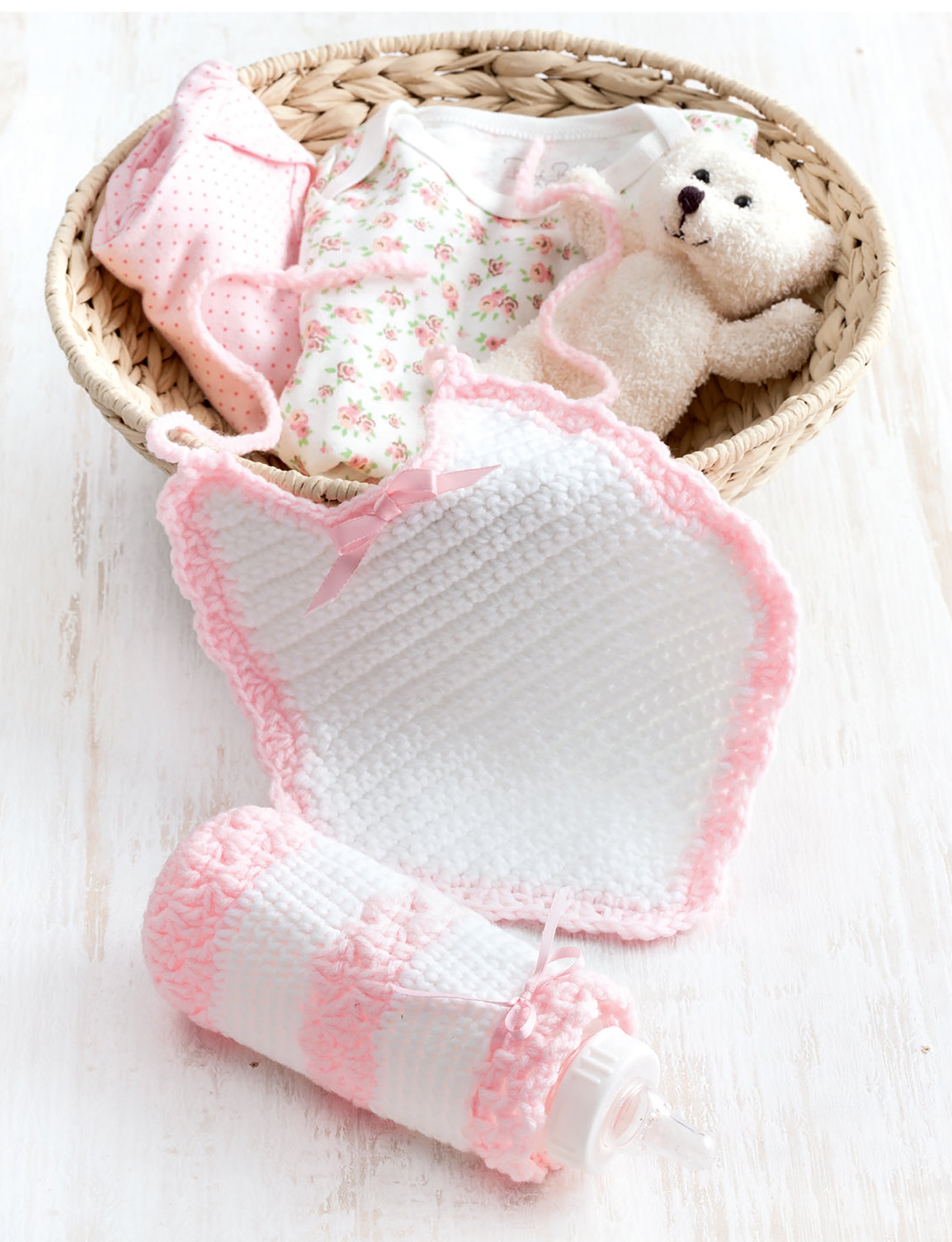 A wicker basket displays Leisure Arts Bibs, Bottle Covers & Booties with a small teddy bear, all set on a white wooden surface.