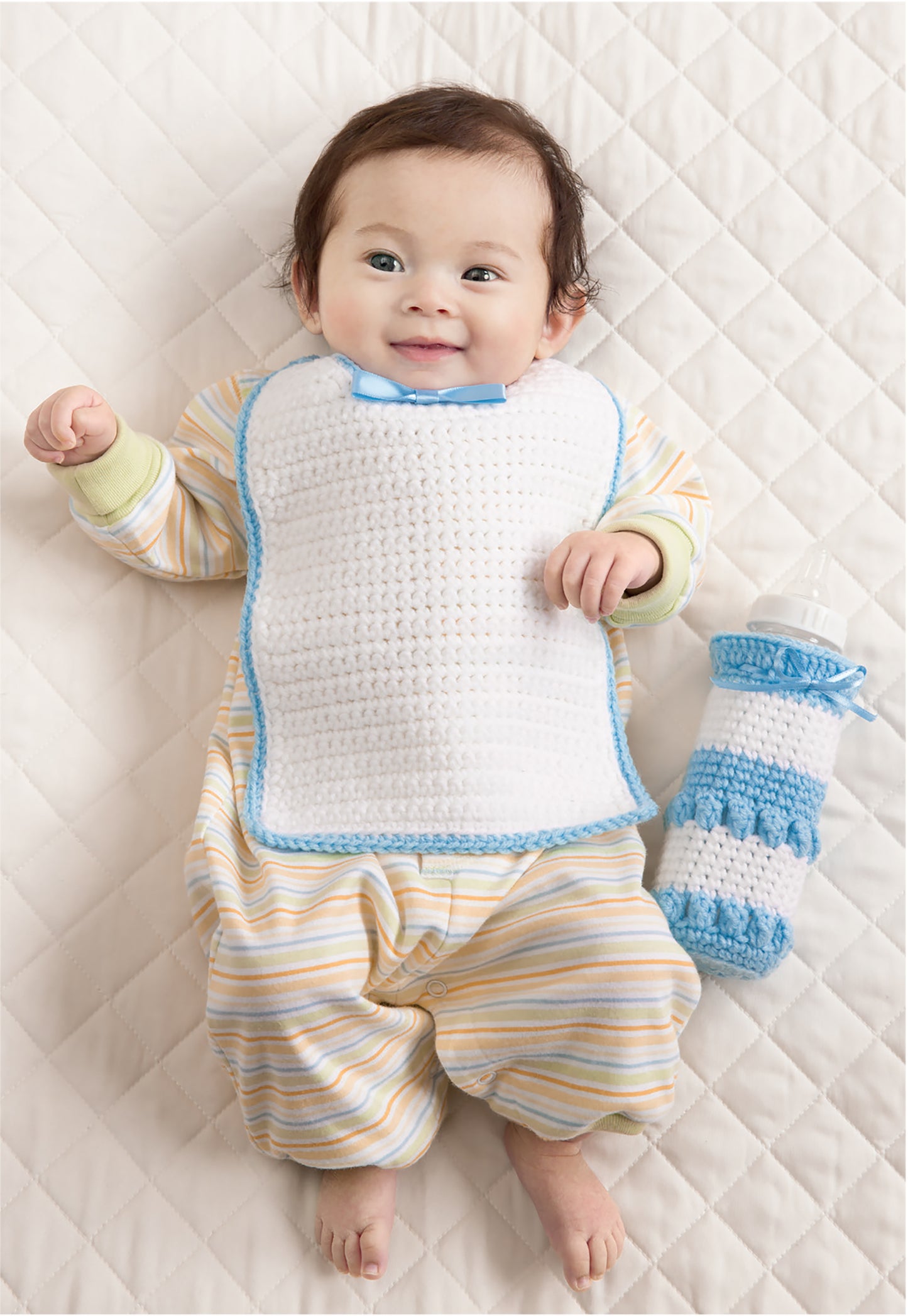 A smiling baby enjoys comfort with Leisure Arts Bibs, Bottle Covers & Booties, resting on a quilted blanket while wearing striped pajamas and a crocheted bib, with cozy booties and a blue-and-white bottle cover nearby.