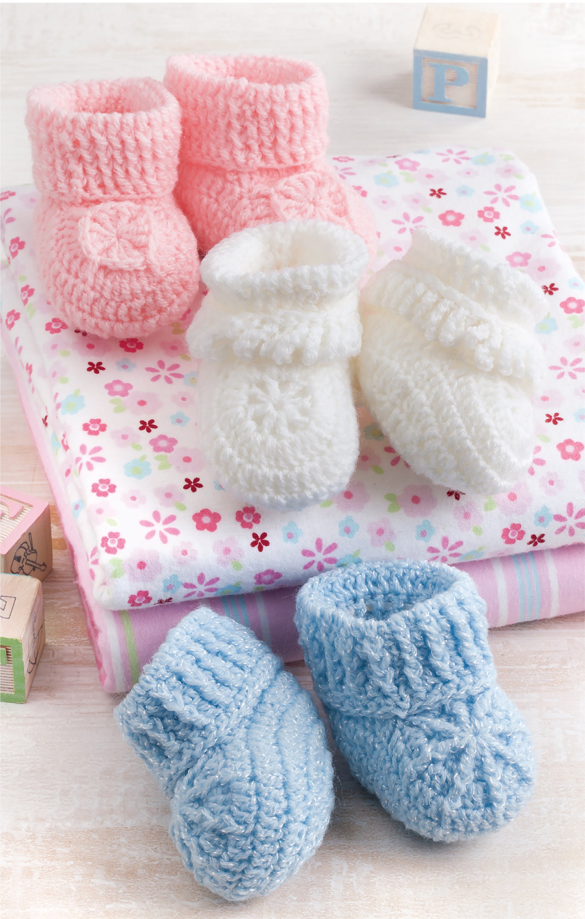 Leisure Arts Bibs, Bottle Covers & Booties set features pink, white, and blue knitted booties on a floral-patterned blanket, with matching bottle covers and wooden alphabet blocks in the background.