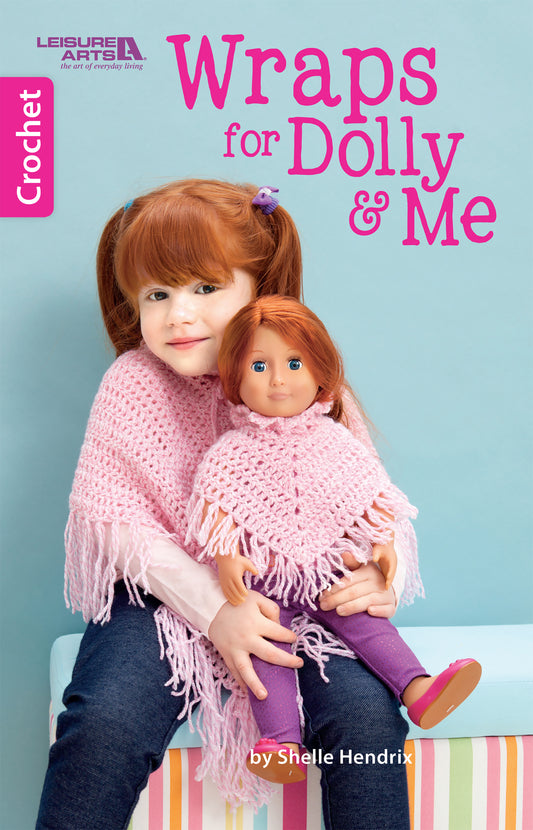 A young red-haired girl with a pink hairband holds a doll; both wear matching pink crocheted shawls made from beginner patterns in the book "Crochet Wraps for Dolly & Me" by Leisure Arts. The title and brand appear in bold pink letters.