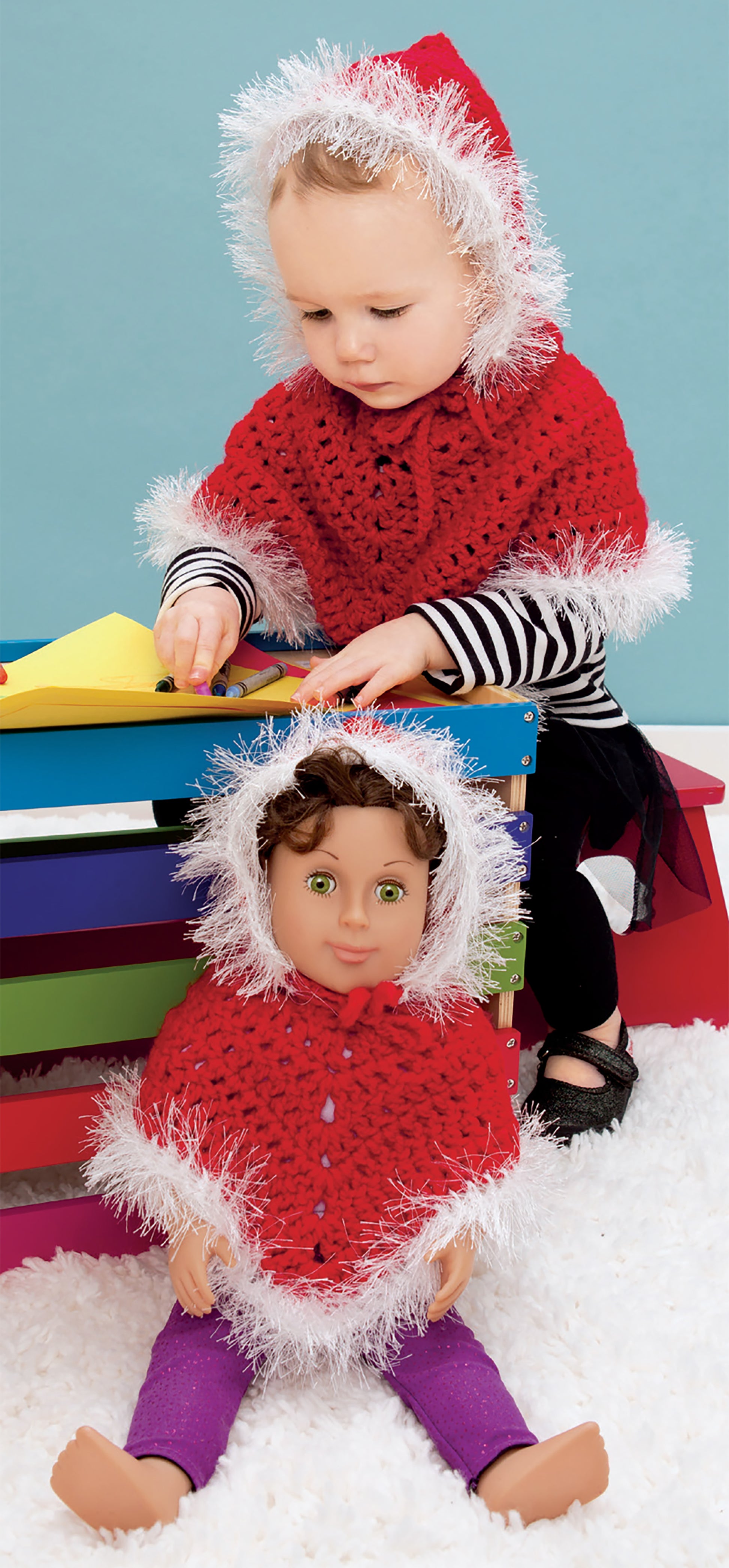 A toddler and doll, both in red Crochet Wraps for Dolly & Me by Leisure Arts, play—the toddler at a colorful table with paper, the doll on a white rug in front of the table.