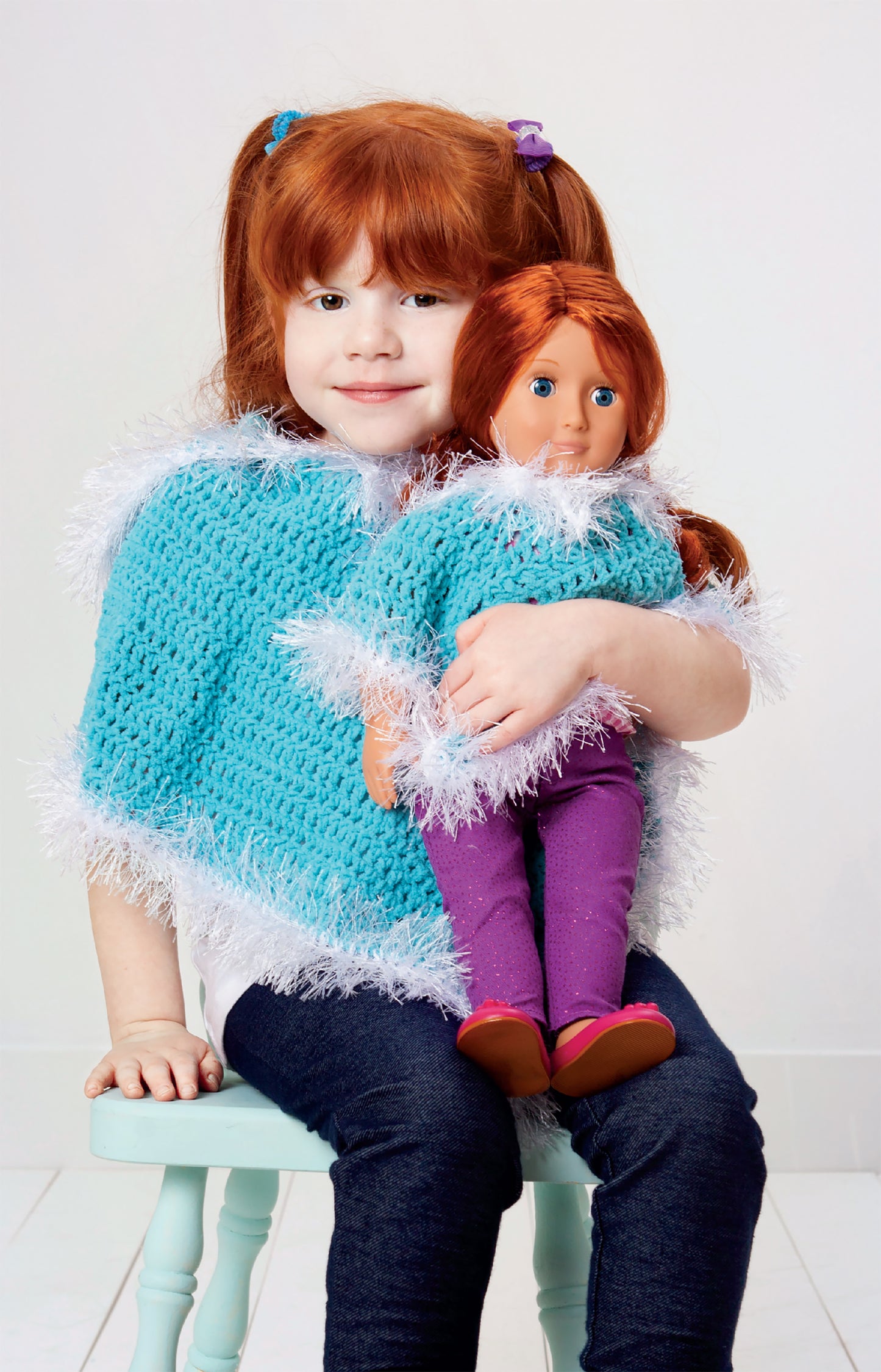 A young red-haired girl sits smiling on a chair, holding her doll; both wear matching blue and white fuzzy wraps made with Leisure Arts’ Crochet Wraps for Dolly & Me patterns.