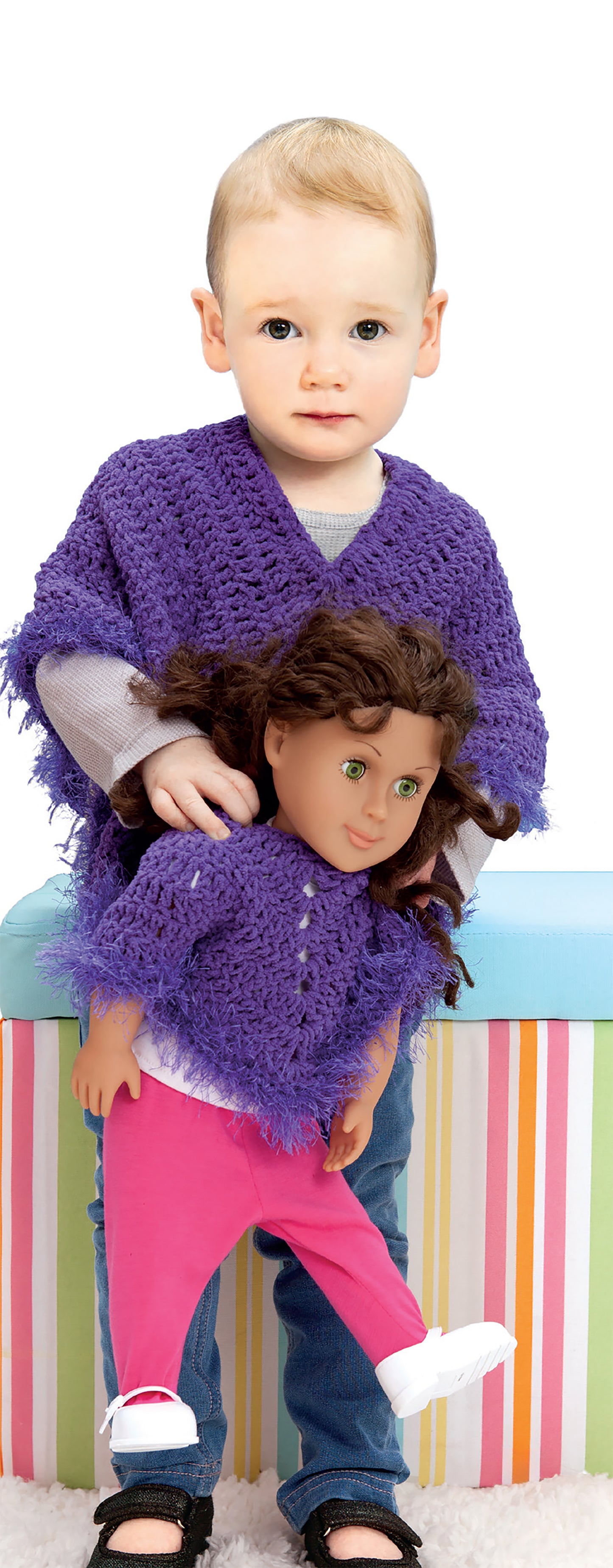A young child wears a purple sweater and holds a doll in a matching one, both made from Leisure Arts' Crochet Wraps for Dolly & Me, with a colorful striped box in the background.