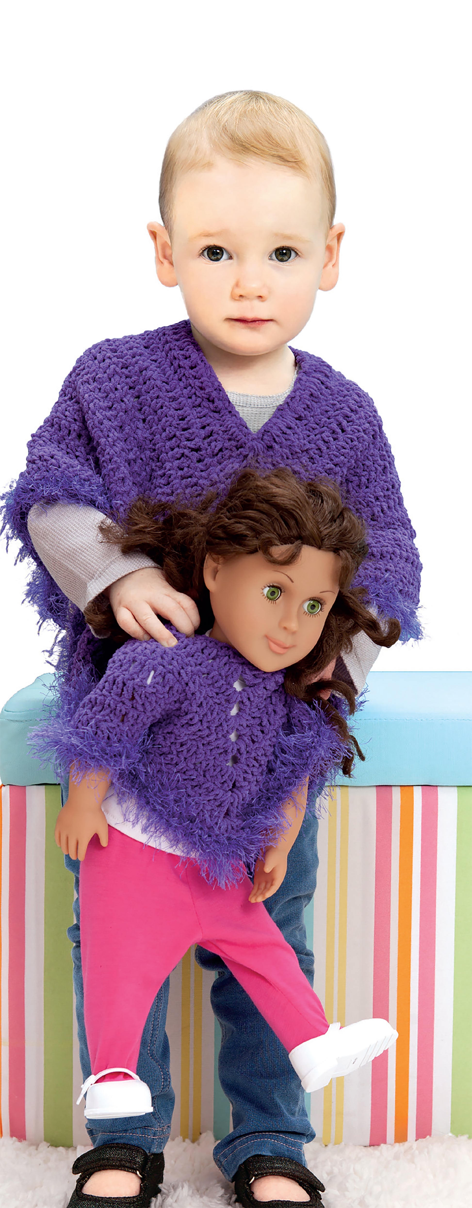 A young child wears a purple sweater and holds a doll in a matching one, both made from Leisure Arts' Crochet Wraps for Dolly & Me, with a colorful striped box in the background.