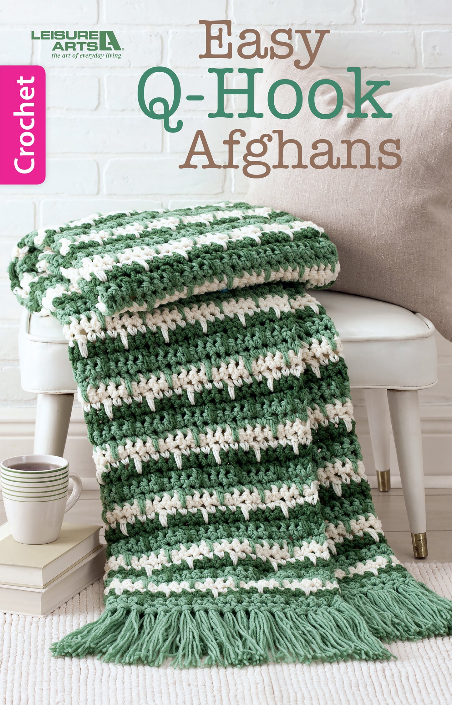 A green and white striped afghan made with a jumbo Q hook from Leisure Arts' "Easy Q-Hook Afghans Digital Download" drapes over a white bench. "Crochet" appears on a pink banner, with a white brick wall in the background.