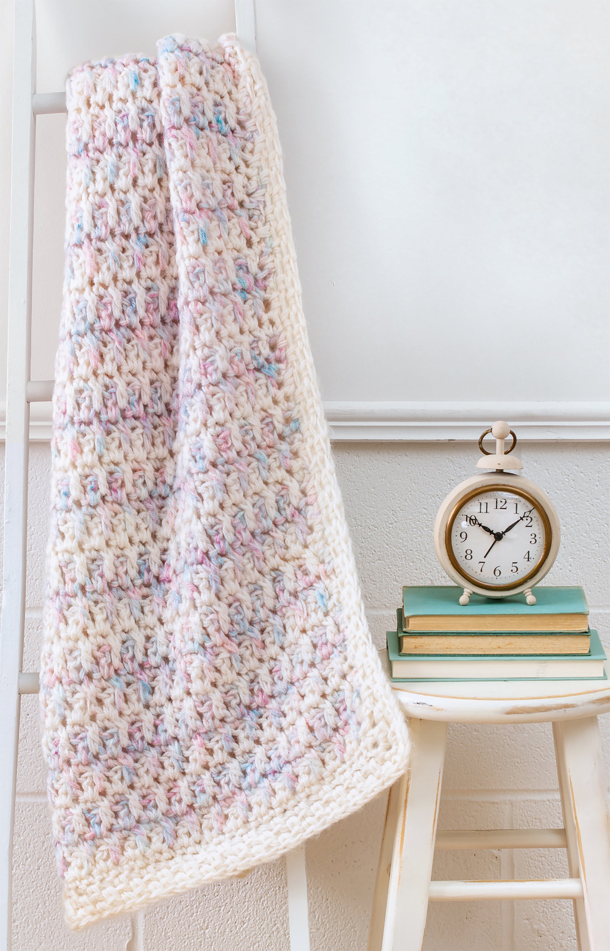 A pastel-speckled afghan, made with the Easy Q-Hook Afghans Digital Download by Leisure Arts, hangs on a ladder beside books and a vintage alarm clock—showcasing an ideal beginner project using a jumbo Q hook.