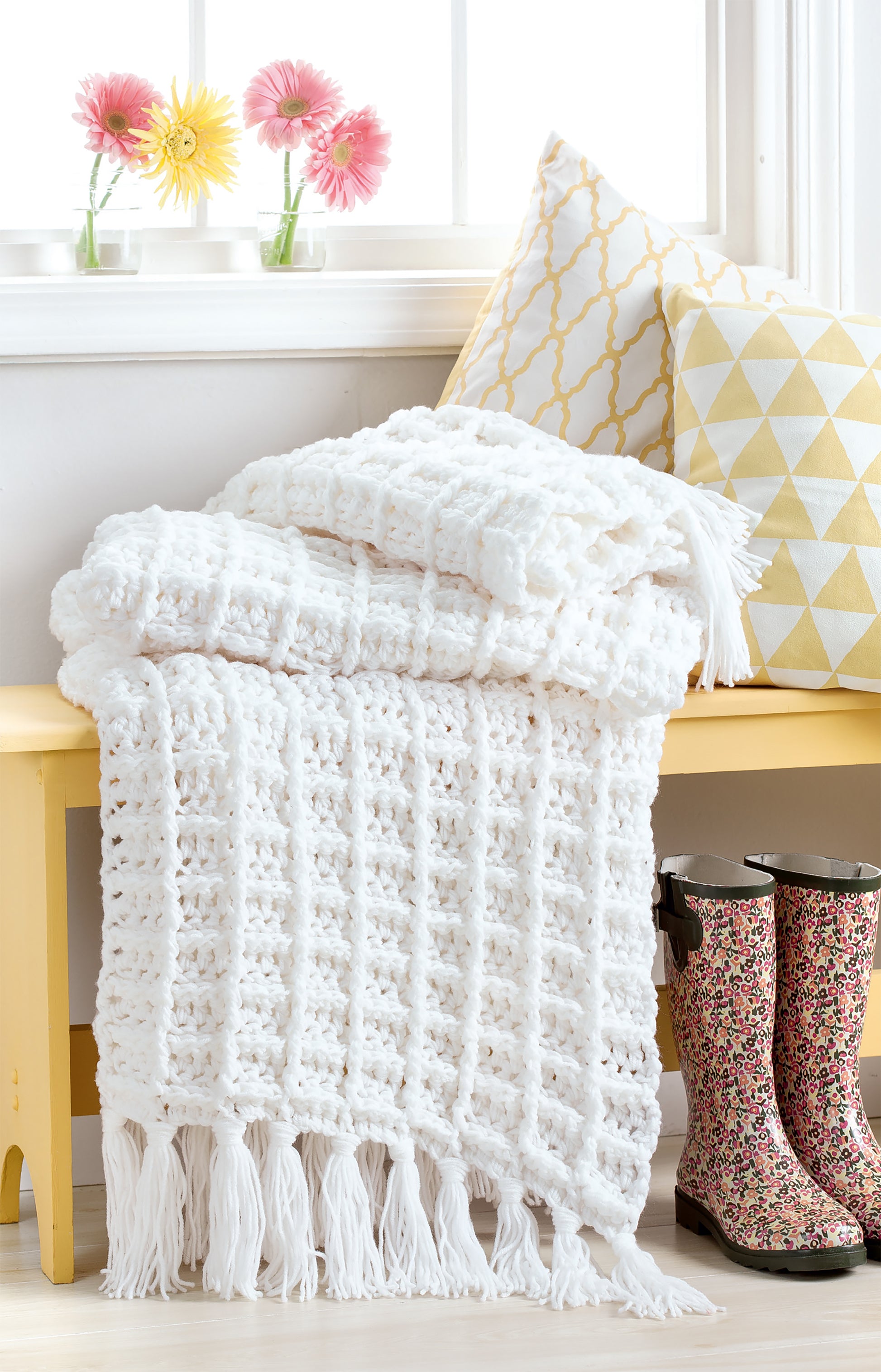 A white crochet-style throw from Leisure Arts’ Easy Q-Hook Afghans (digital download) drapes over a yellow bench with patterned pillows, floral rain boots below, and bright flowers in vases on the windowsill behind.