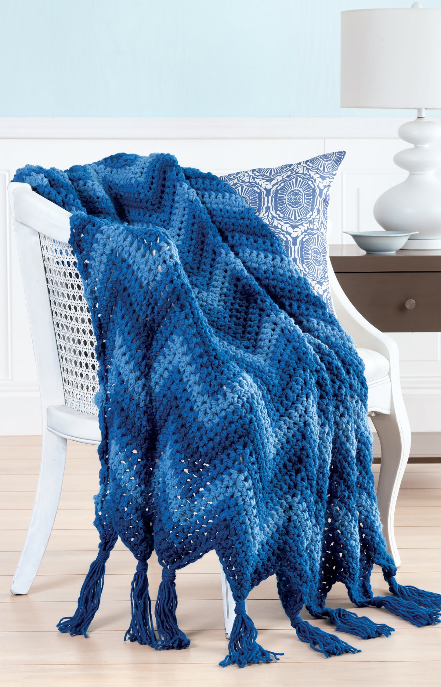 A white chair holds a blue and white pillow and a blue chevron afghan, crocheted with Leisure Arts' Easy Q-Hook Afghans Digital Download. The chunky design uses a jumbo Q hook. A white lamp and brown table appear in the background.