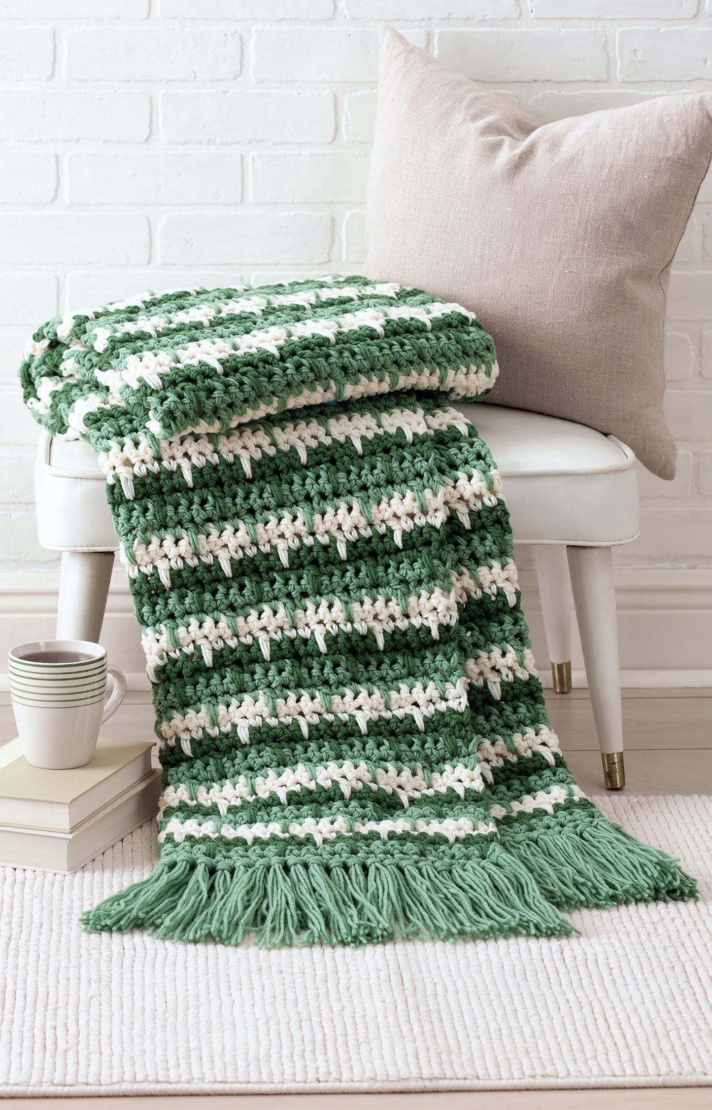 The Easy Q-Hook Afghans Digital Download by Leisure Arts features a green and white striped crochet afghan with fringe, draped over a white stool near a beige pillow, stacked books, and cups on a light rug against a white brick wall.