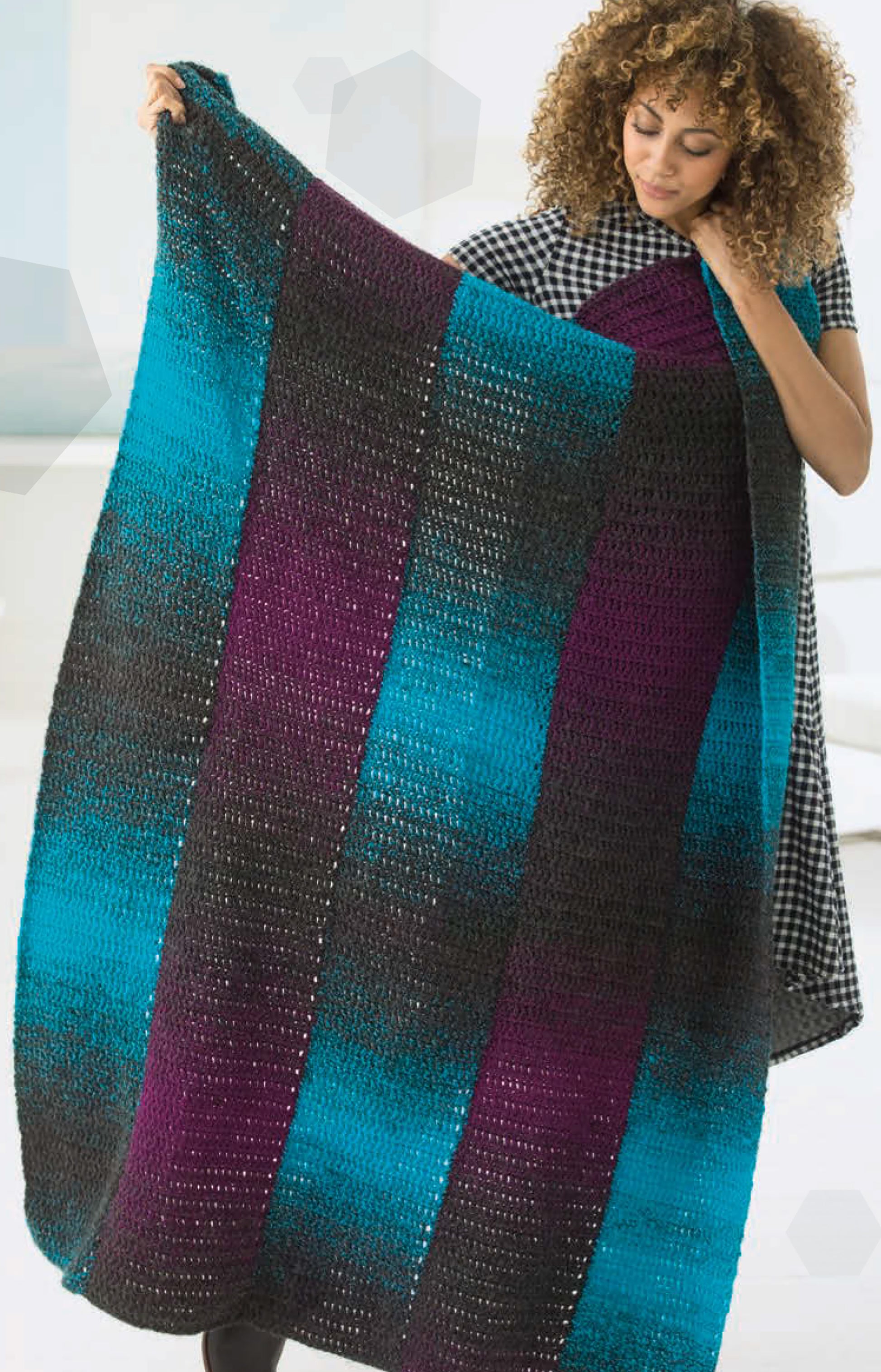 A woman with curly hair holds up a large crocheted blanket made with Leisure Arts Simply Scarfie Crochet, featuring bold black, teal, and purple stripes—a perfect inspiration for cozy crochet projects.