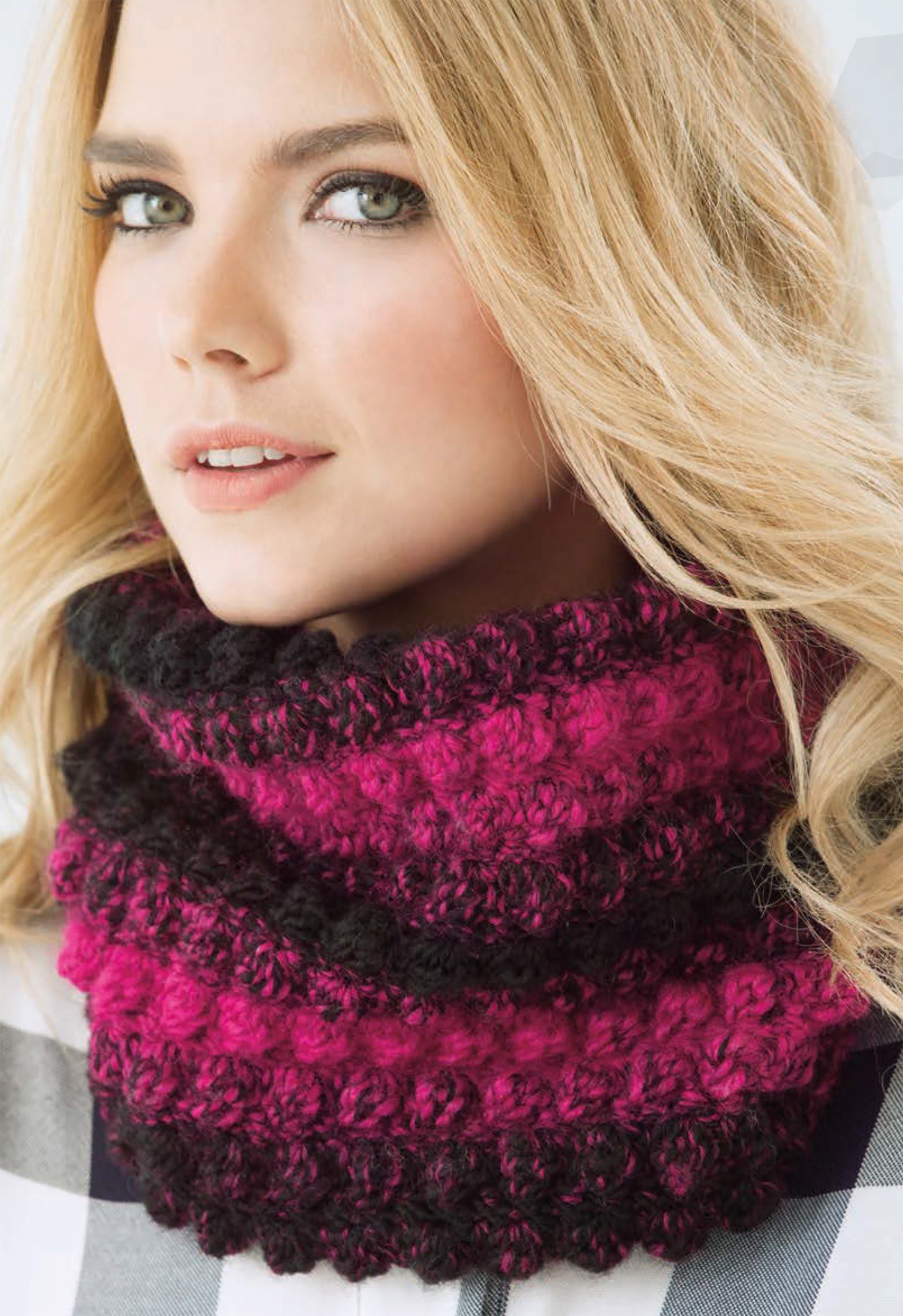 A woman with long blonde hair models a thick, textured cowl from the Simply Scarfie Crochet collection by Leisure Arts. Made with deep pink and black yarn, she pairs it with a plaid shirt—perfect inspiration for your next crochet project.