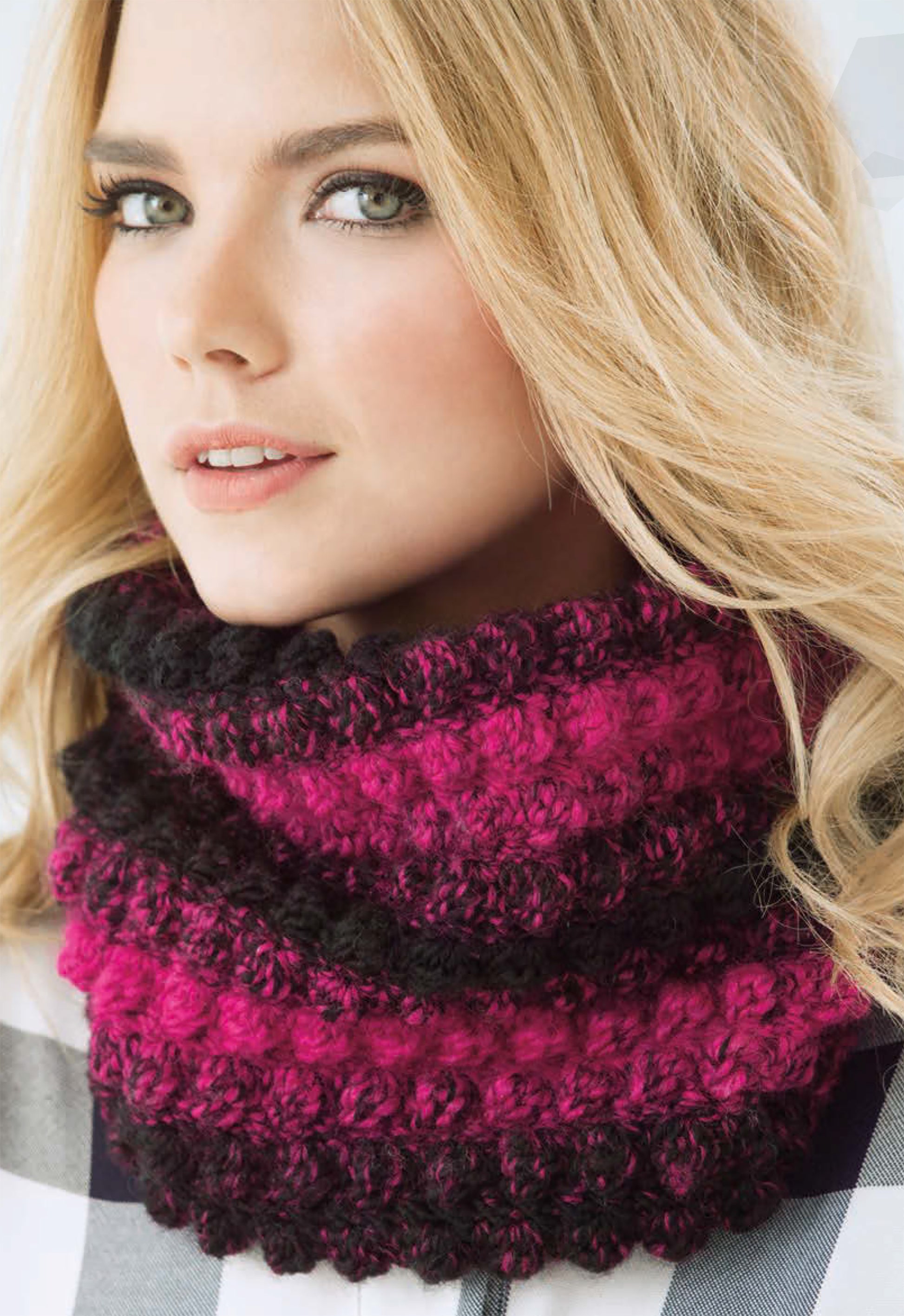 A woman with long blonde hair models a thick, textured cowl from the Simply Scarfie Crochet collection by Leisure Arts. Made with deep pink and black yarn, she pairs it with a plaid shirt—perfect inspiration for your next crochet project.